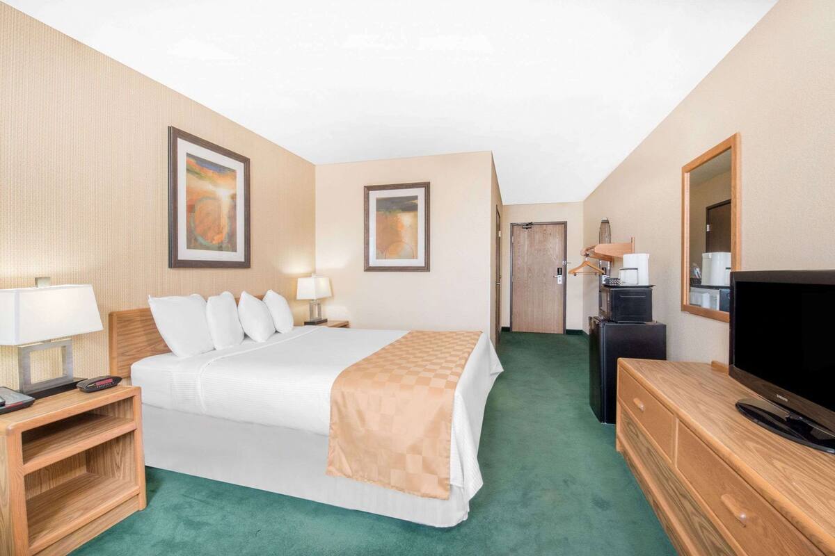 Een hotelkamer met een bed, nachtkastje, lamp, televisie, kapstok en een deur naar een andere kamer.