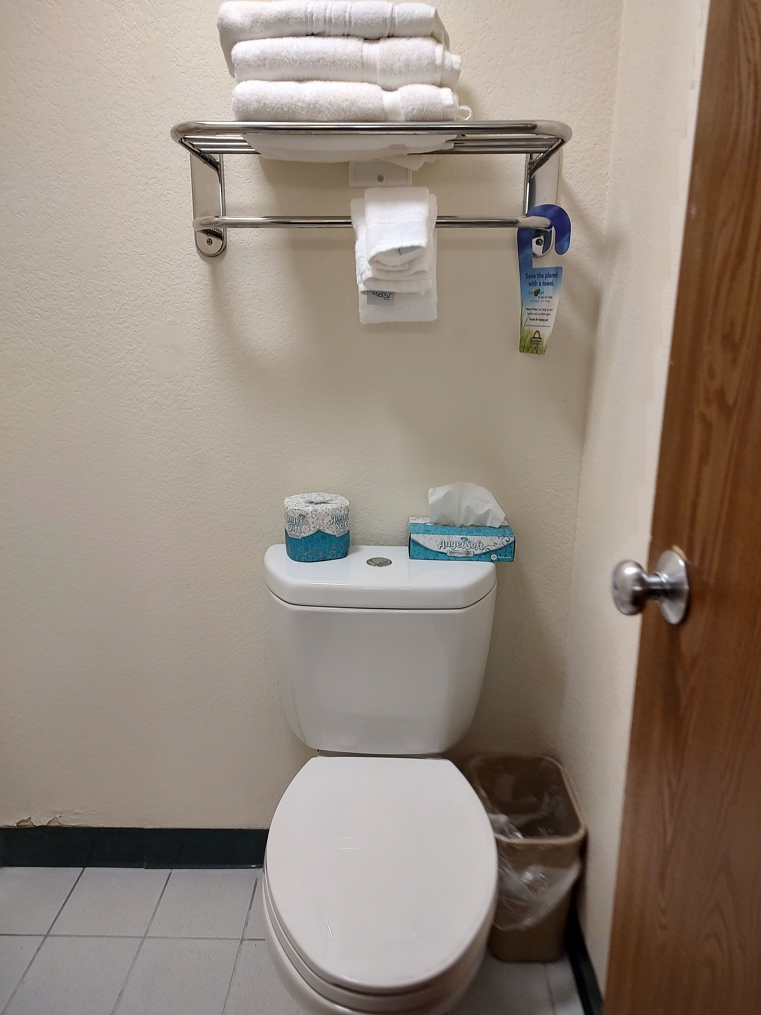 A bathroom with a toilet, a towel rack with folded towels, toilet paper, and a trash bin.