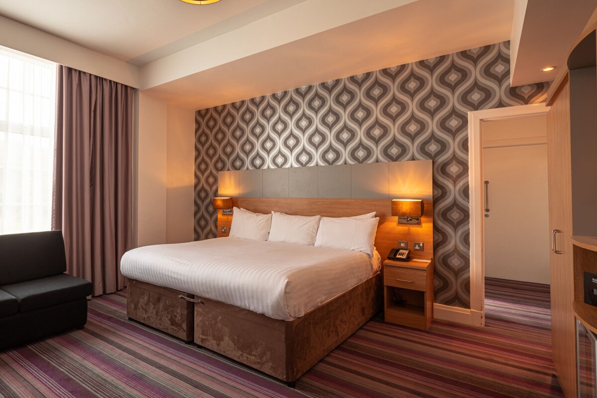 A hotel room with a large bed, a sofa, a patterned wall, and a window with curtains.