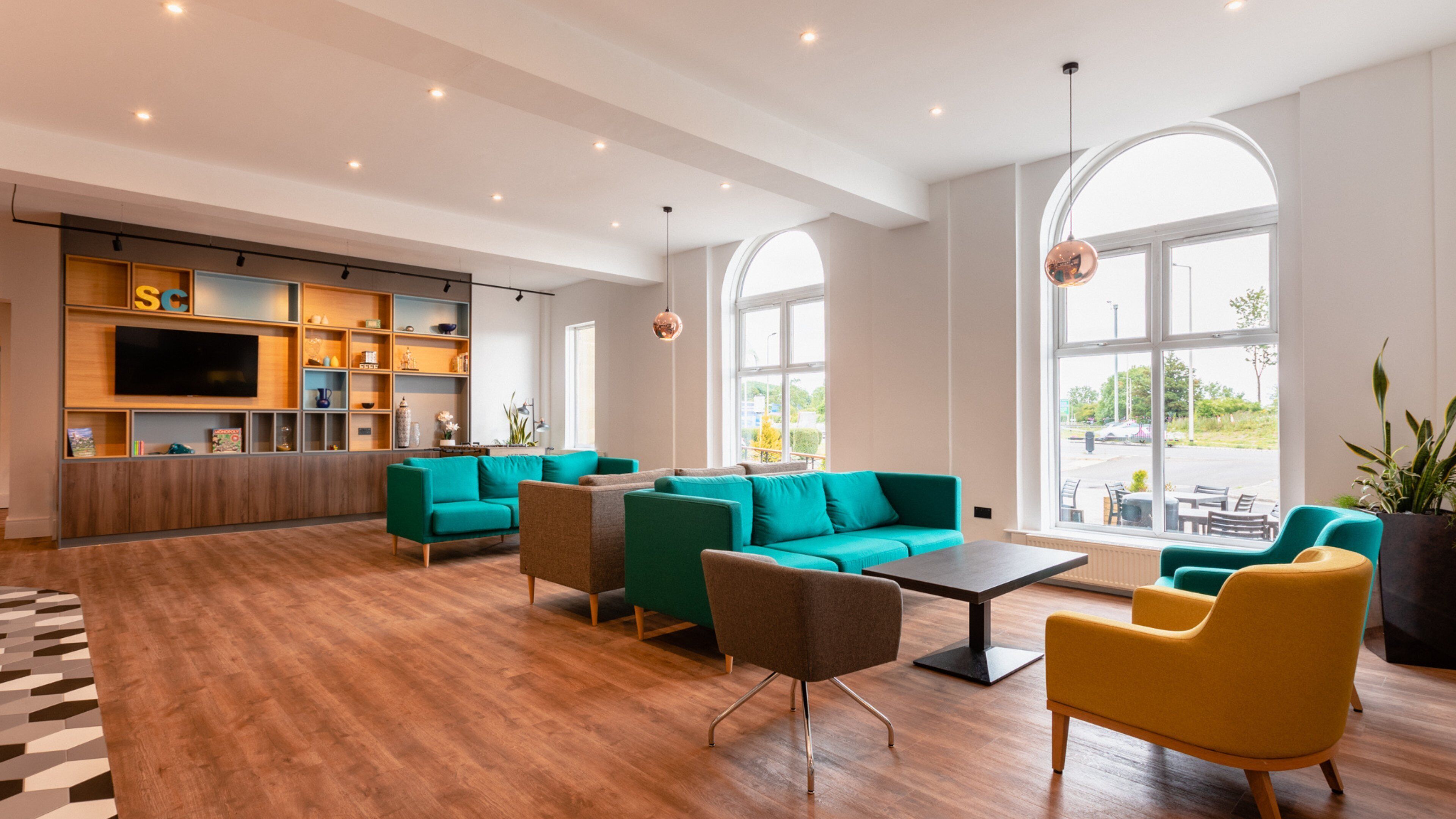A spacious lounge area with teal and yellow seating, a wooden floor, and a television on the wall.
