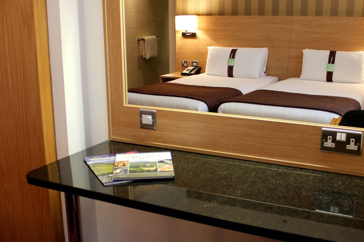 A hotel room with a glass desk, a wooden chair, a bed with white linens, a mirror, and a lamp.