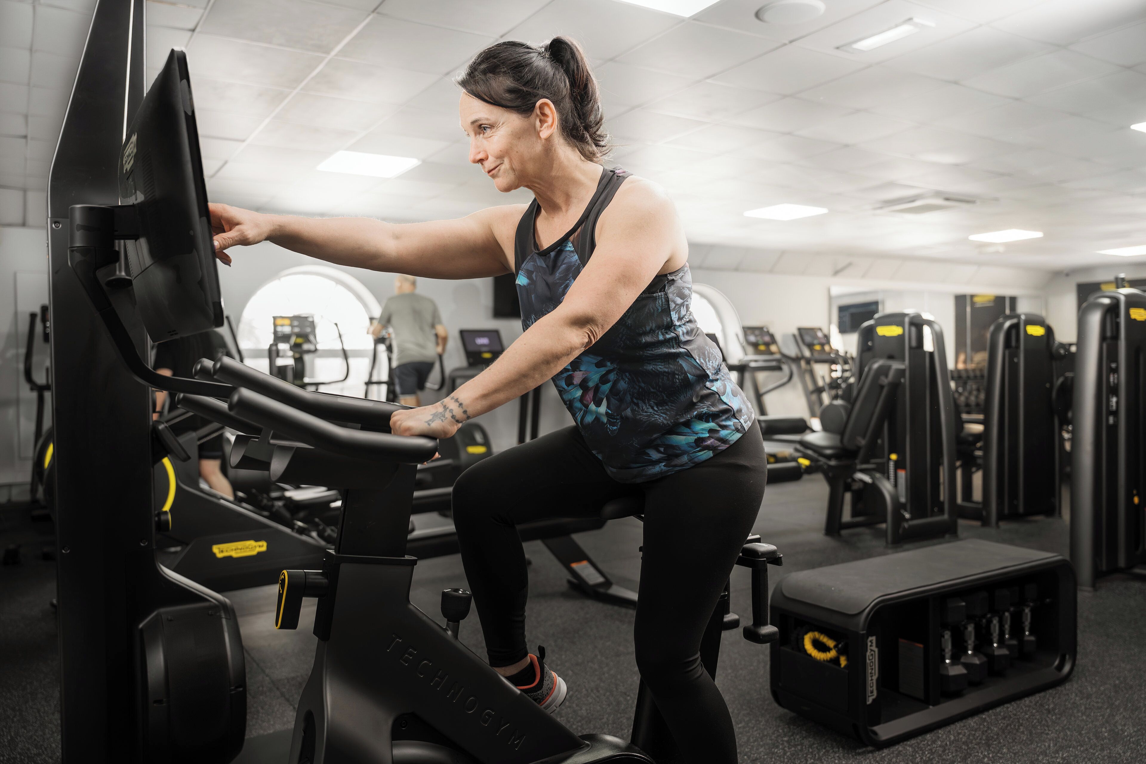 Een vrouw die een hometrainer gebruikt in een sportschool, met diverse fitnessapparaten op de achtergrond.