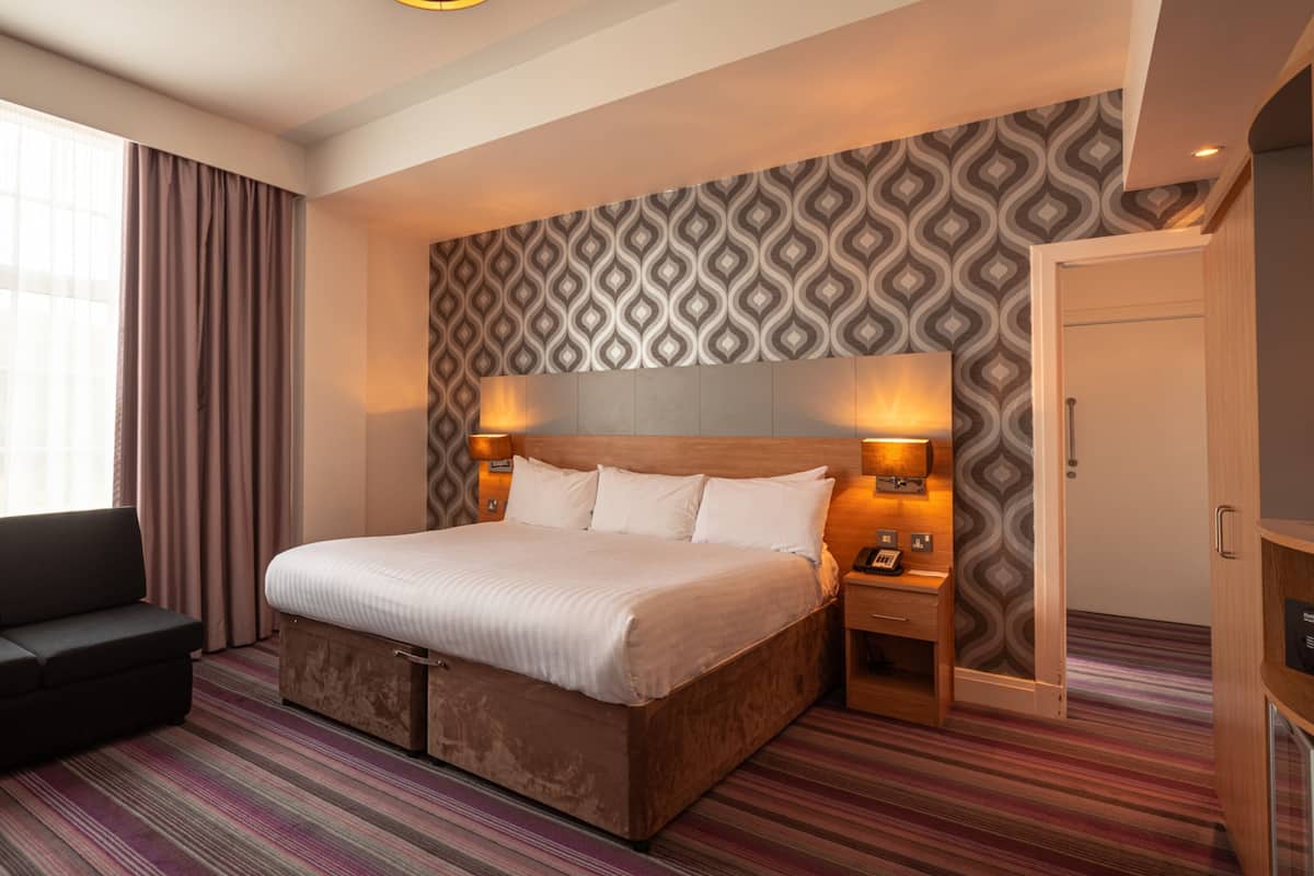 A hotel room with a large bed, a sofa, a patterned wall, and a window with curtains.