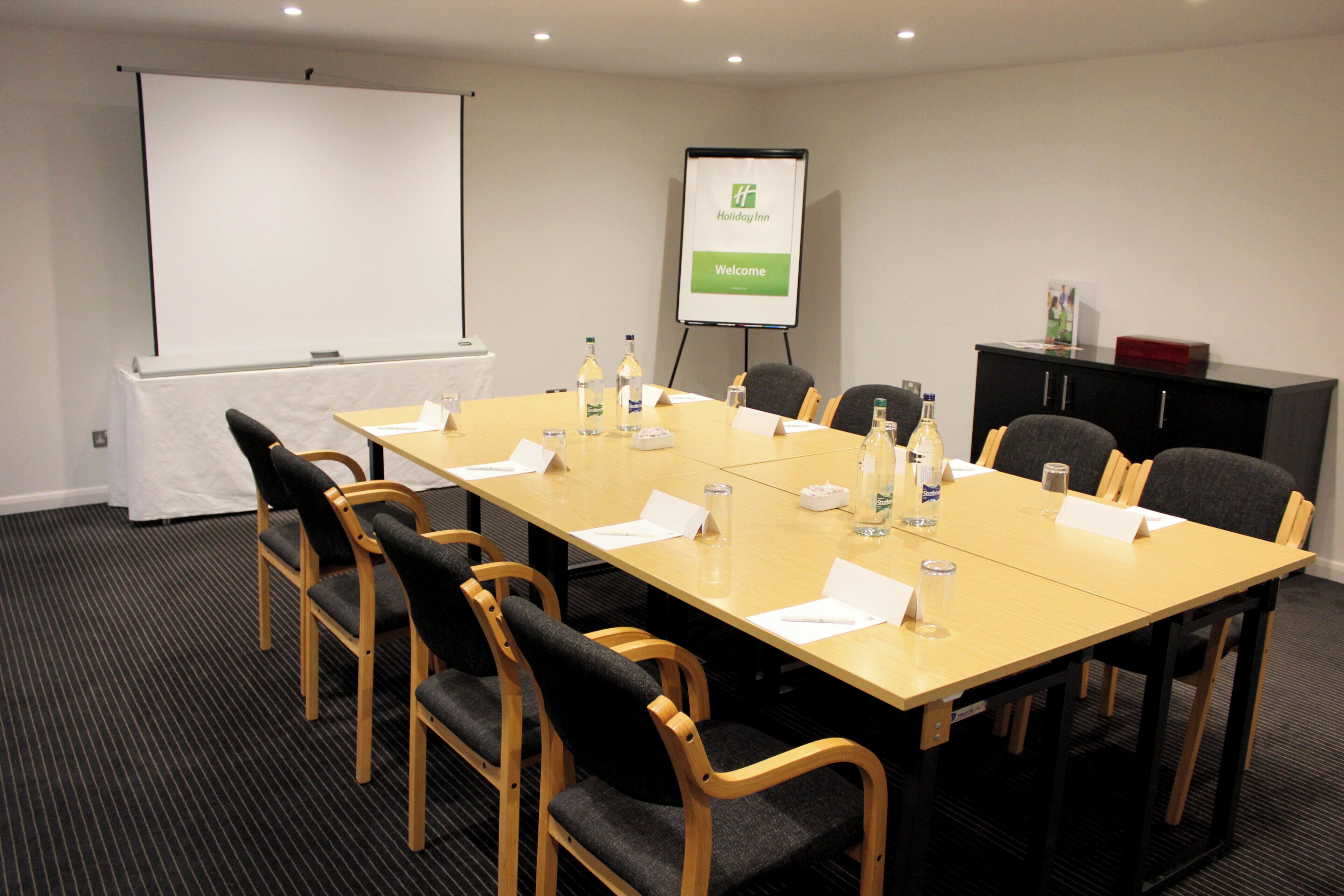 Een vergaderruimte met een lange tafel, stoelen, een scherm voor een beamer, een whiteboard en een bord met een welkomstbericht.