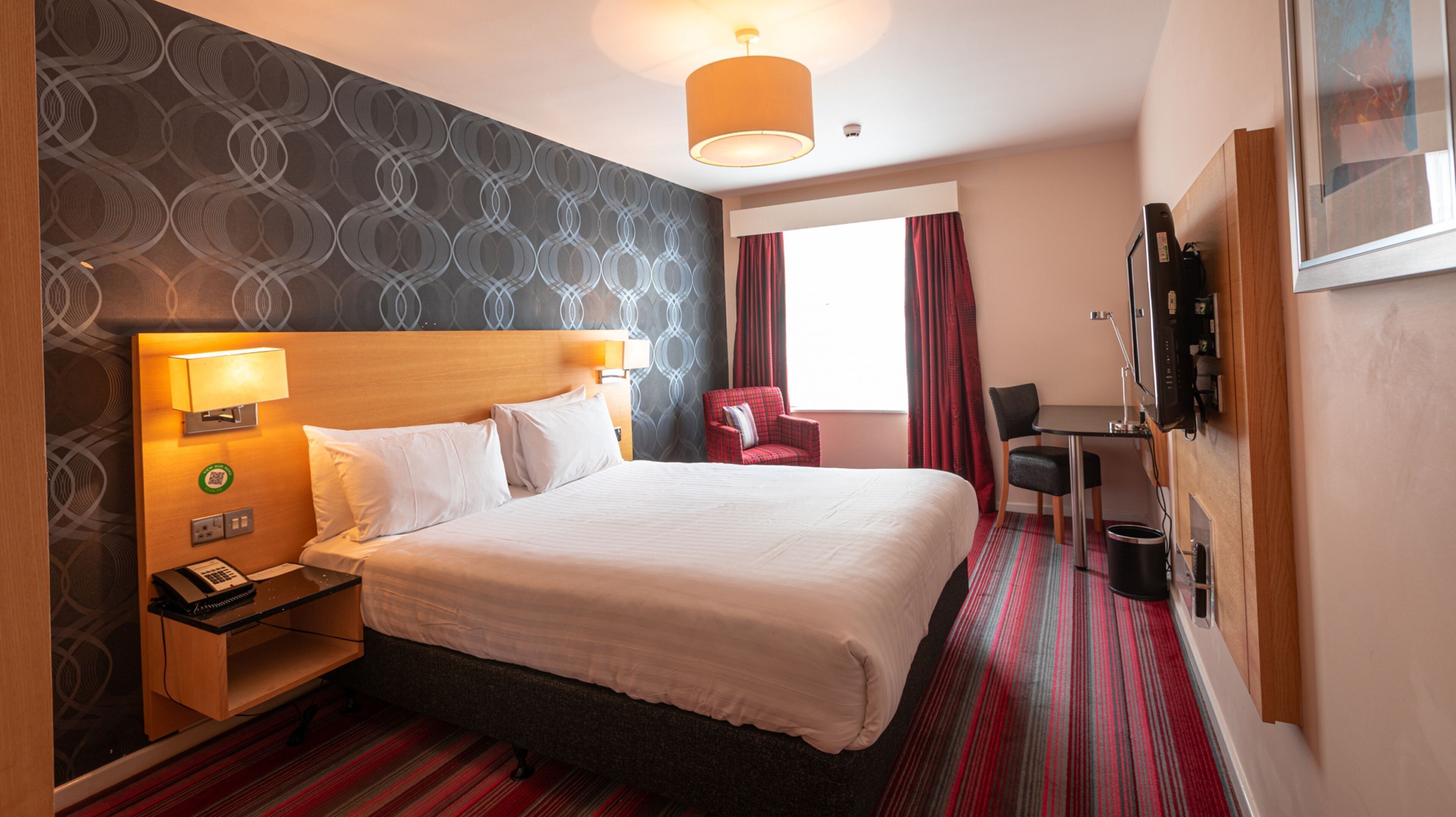 A hotel room with a large bed, a desk, a chair, a TV, and a window with curtains.