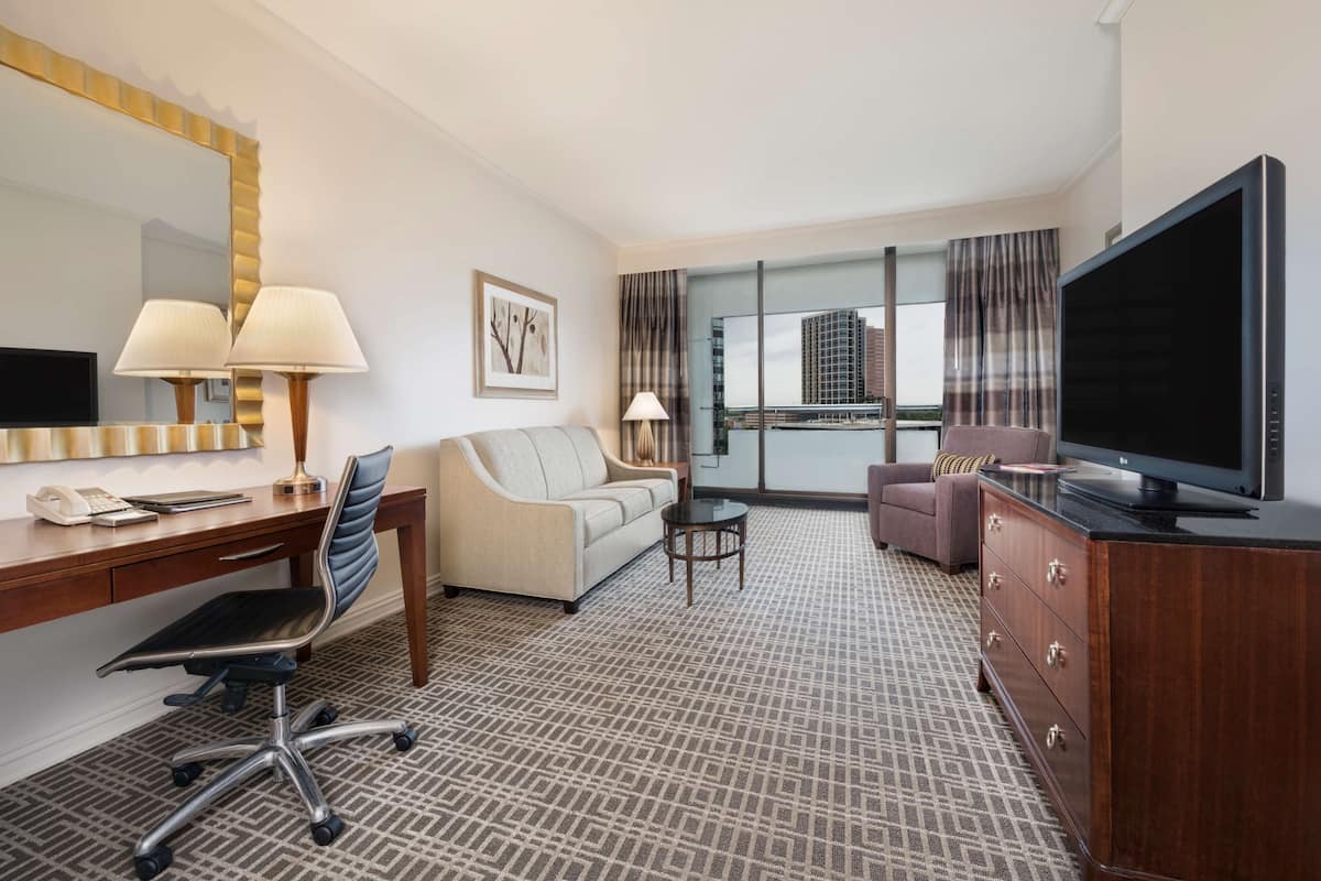 A hotel room with a desk, office chair, sofa, TV, and a large window with a city view.