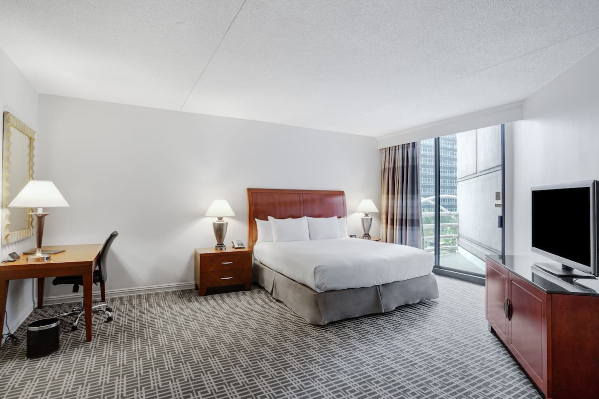 A hotel room with a bed, desk, chair, TV, and large windows.