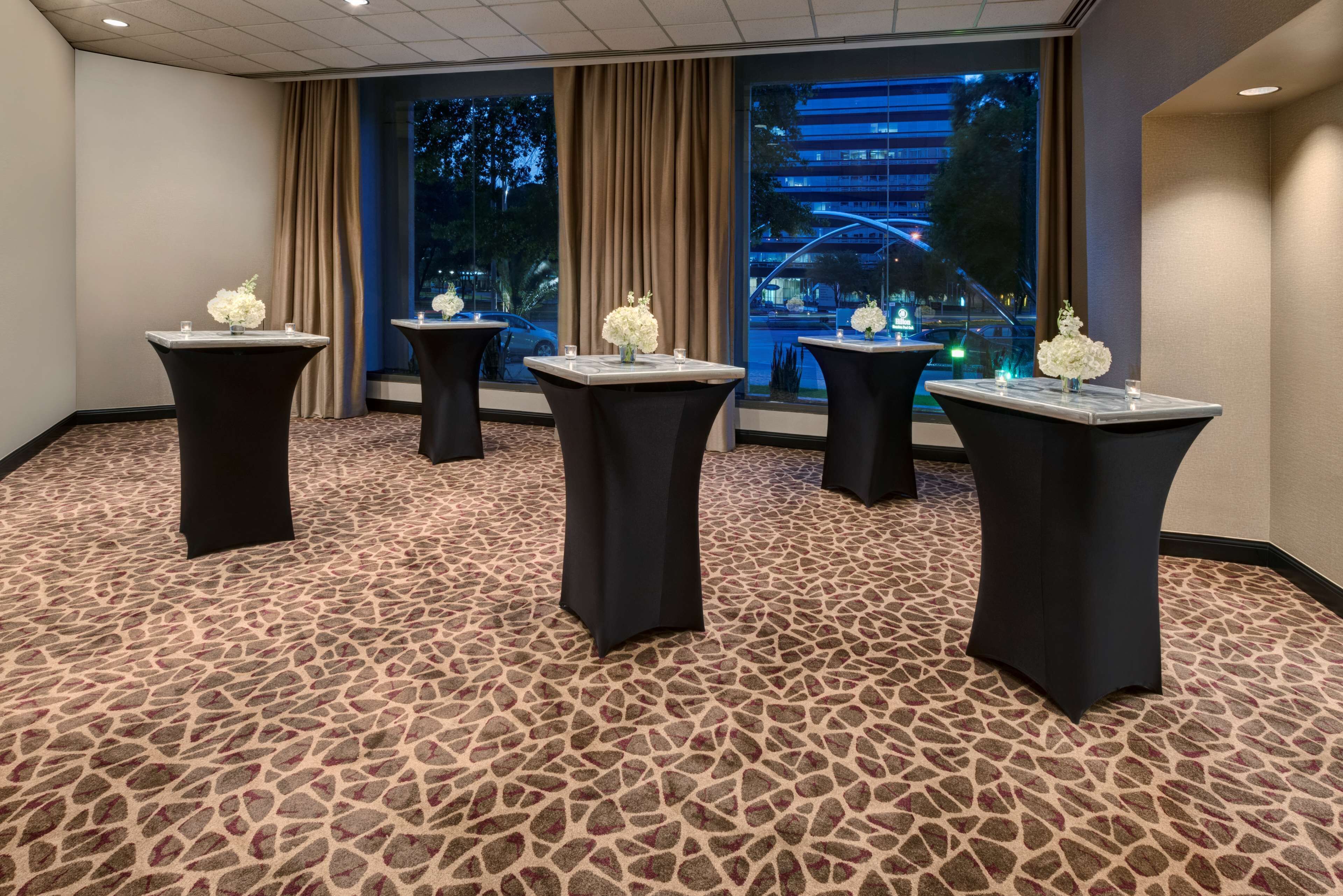 A conference room with four tables, each with a vase of white flowers and candles, set against a patterned carpet and large windows with curtains.