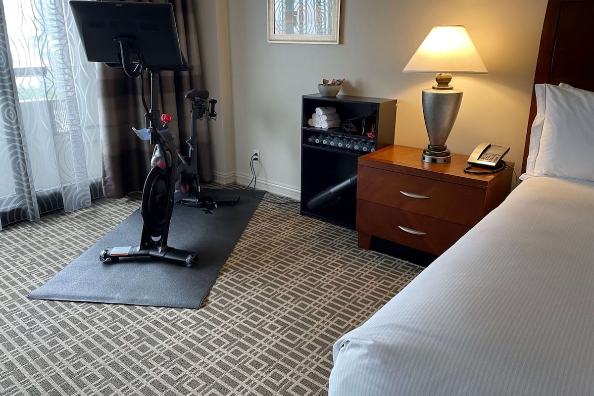 A hotel room with a bed, a desk with a lamp, a phone, a treadmill, and a TV on a stand.