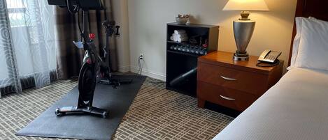 A hotel room with a bed, a desk with a lamp, a phone, a treadmill, and a TV on a stand.