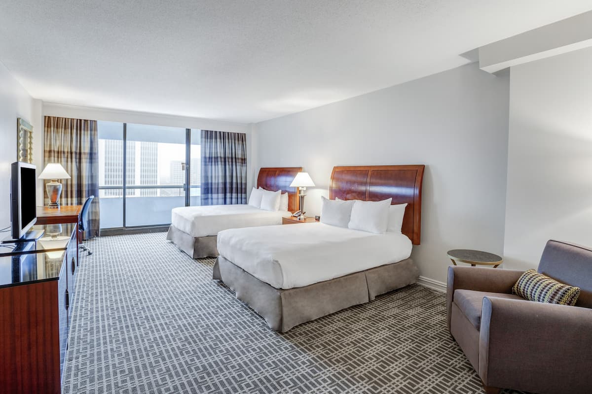 A hotel room with two beds, a desk with a TV, a sofa, and a large window with curtains.