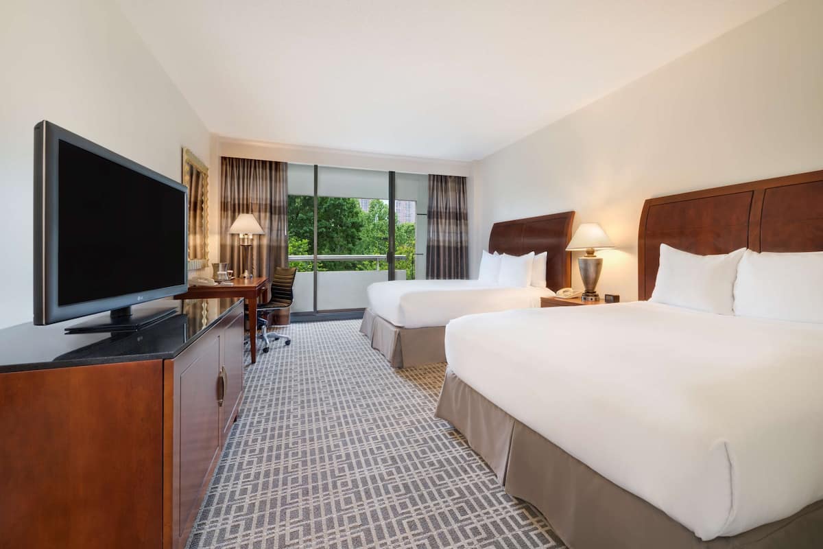 A hotel room with two beds, a TV, a desk with a lamp, and a view of greenery through the window.