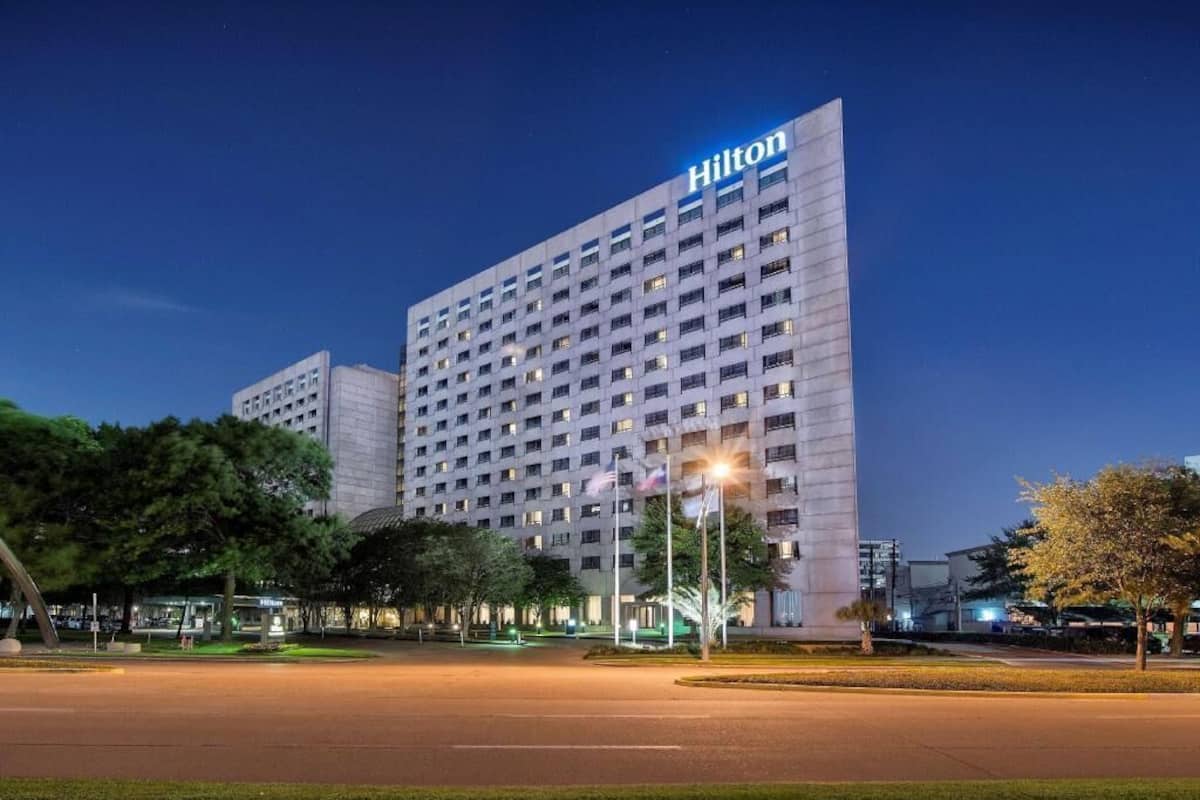 A large, multi-story hotel with a prominent 'Hilton' sign, surrounded by a well-lit parking area and greenery.