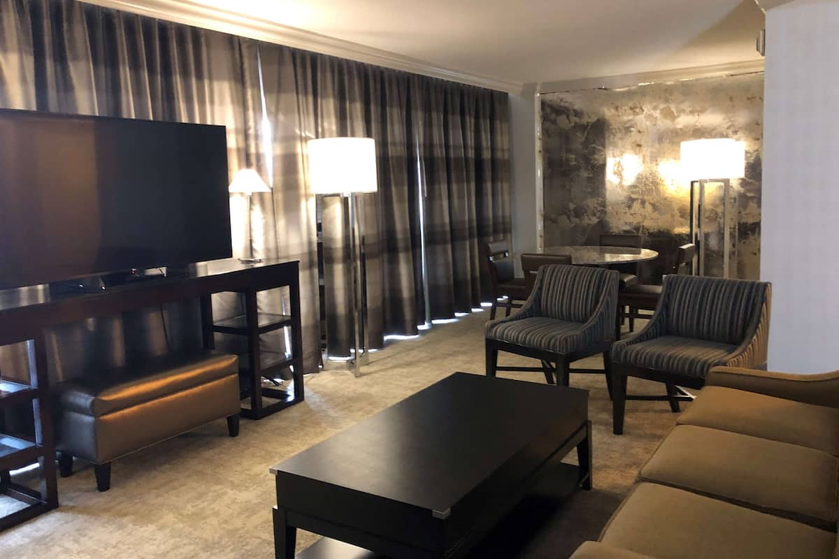 A hotel room with a flat-screen TV, a sofa, a coffee table, a dining table with chairs, and a staircase with curtains.