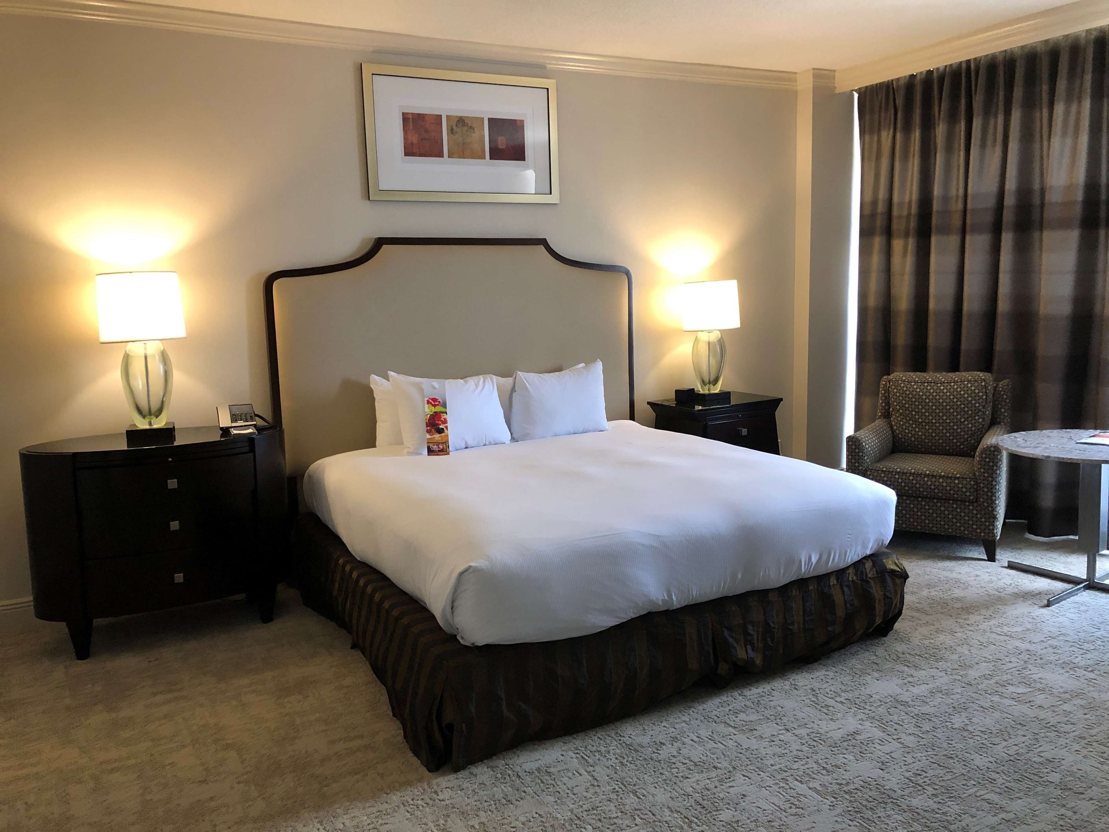 Habitación de hotel con una cama grande, dos lámparas de noche, un cuadro en la pared, una silla y una mesita.