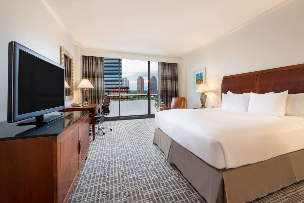 A hotel room with a large bed, a TV on a wooden cabinet, a desk with a chair, and a view of the city through large windows.