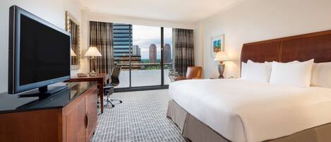 Quarto de hotel com uma cama grande, uma televisão sobre um móvel de madeira, uma escrivaninha com uma cadeira e vista para a cidade através de grandes janelas.