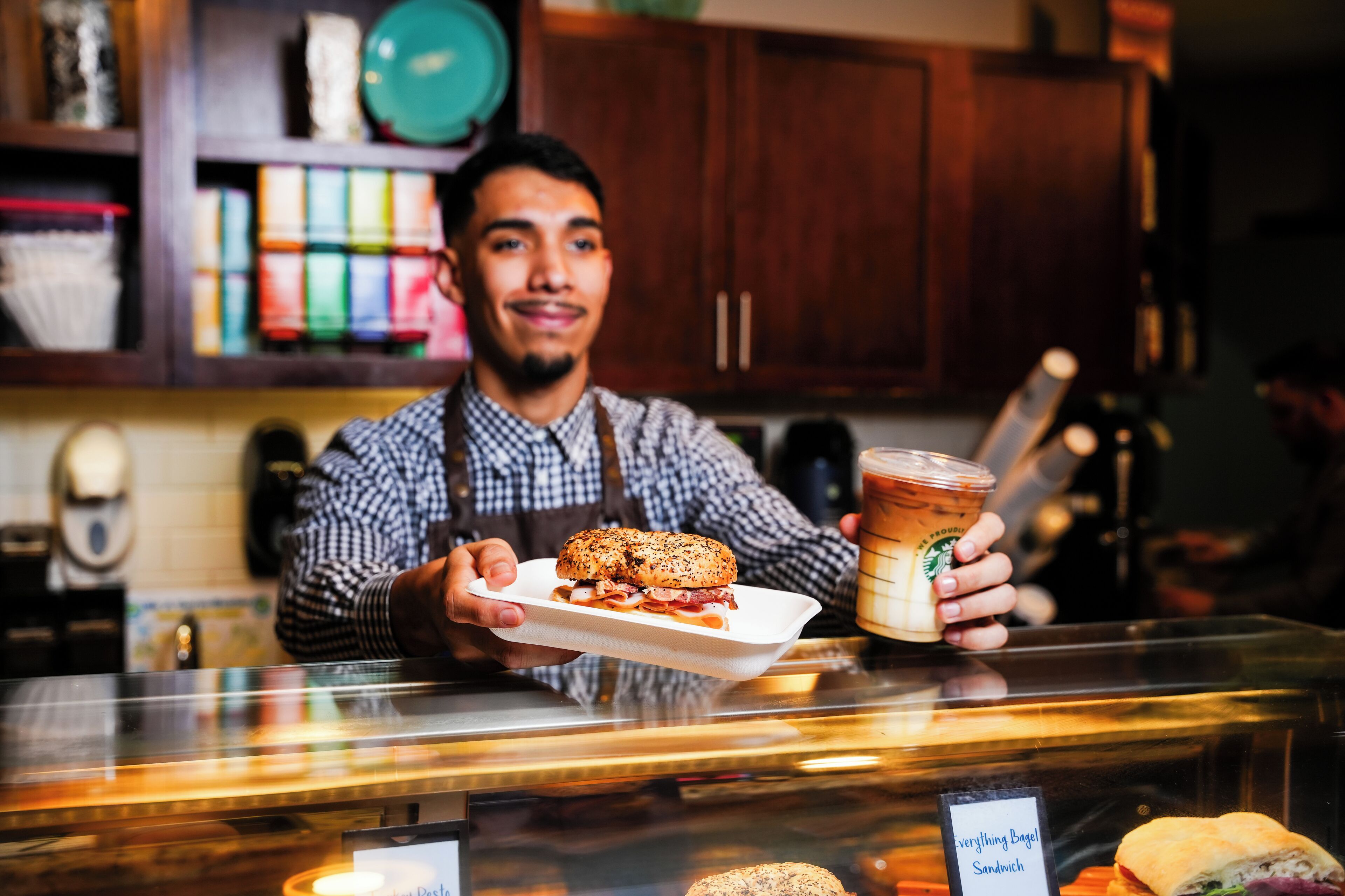 A person holding a sandwich and a drink at a counter with a menu board in the background.