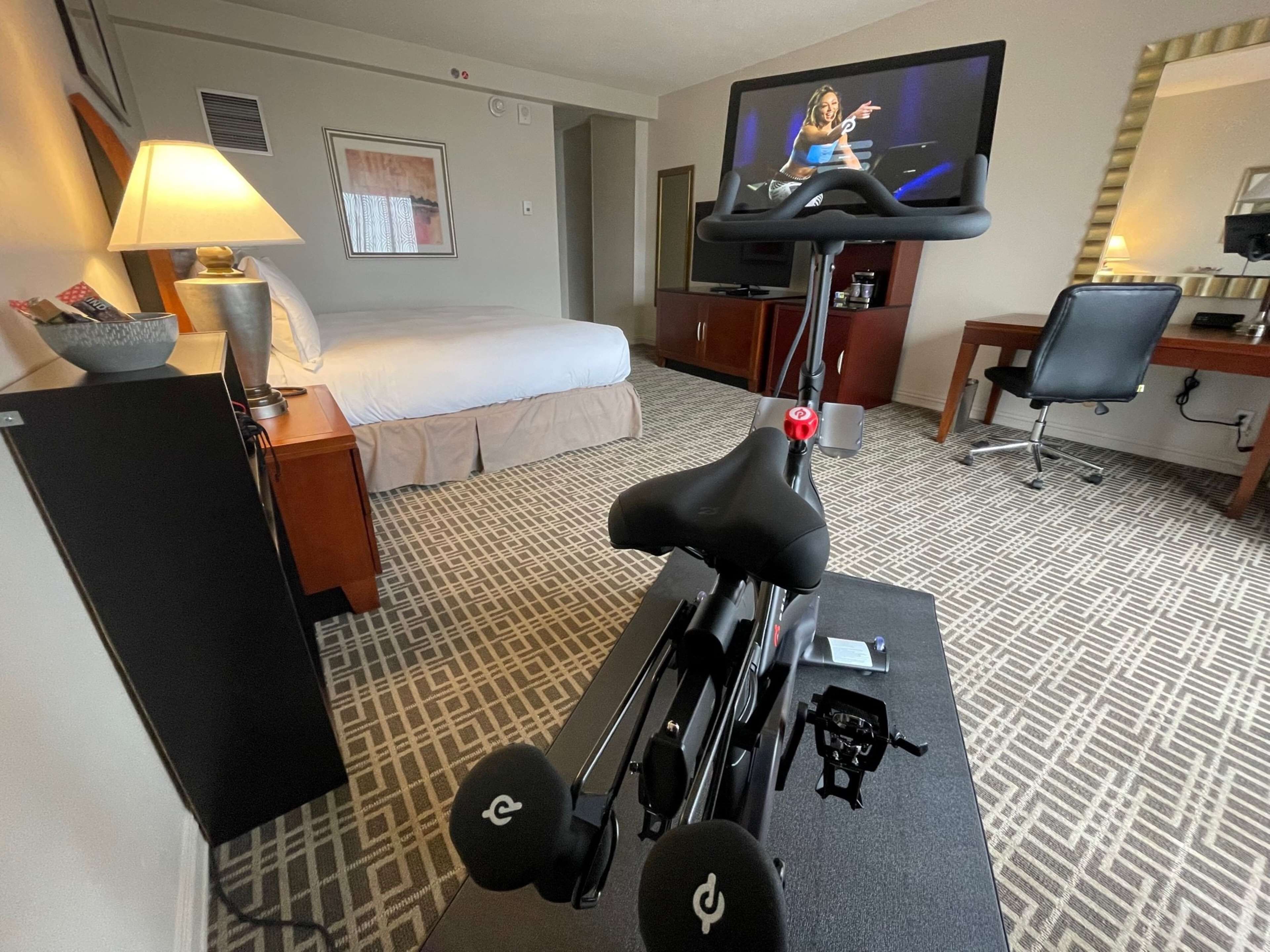 A hotel room with a bed, a desk, a chair, a treadmill, a television, and a lamp.
