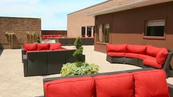 Un patio exterior con mobiliario de mimbre y cojines rojos, una pared de ladrillo y un edificio con ventanas y una puerta.