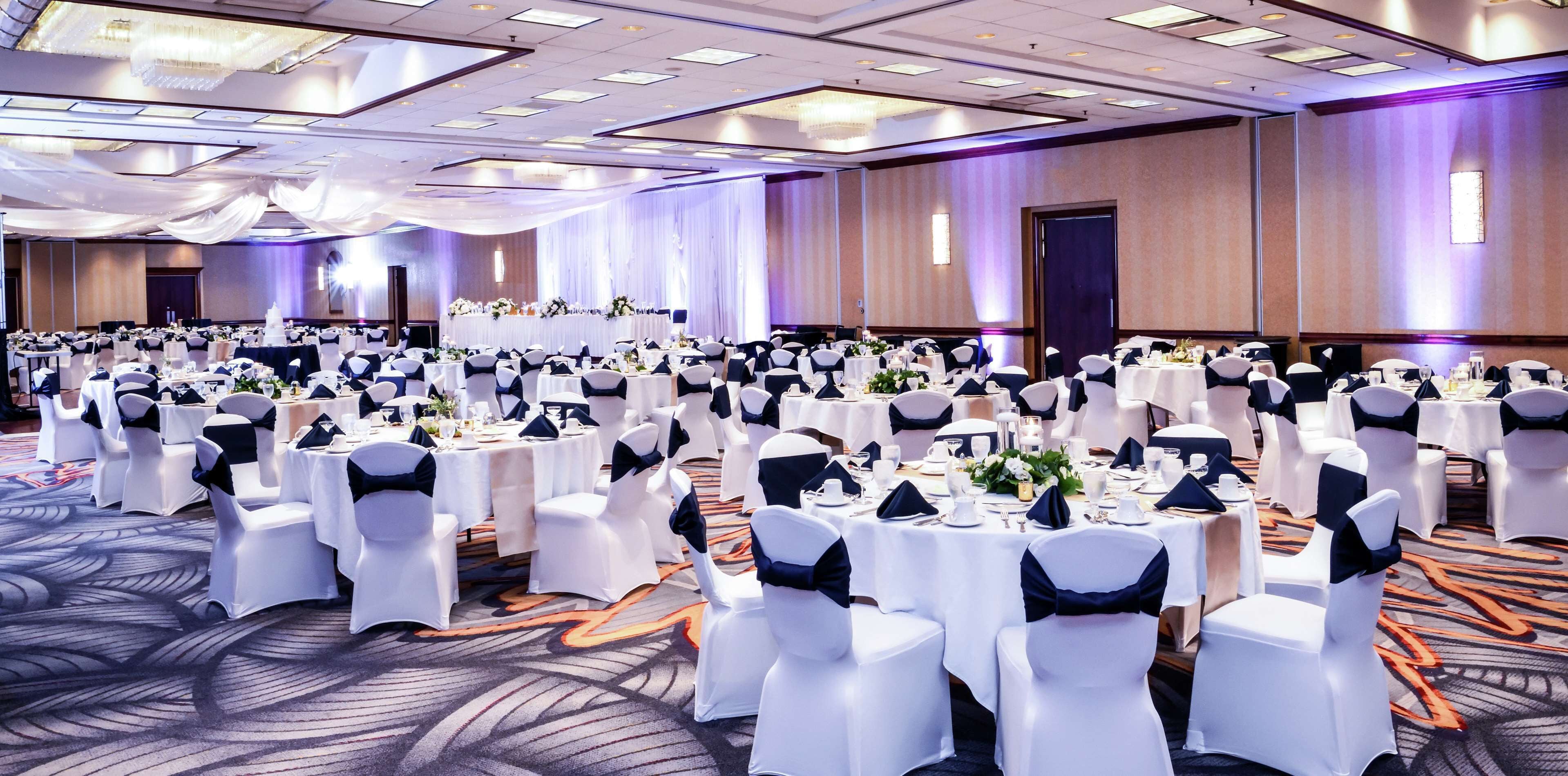 Une vaste salle de banquet, avec des tables rondes dressées pour un événement formel, agrémentées de nappes blanches et de rubans noirs sur les chaises.