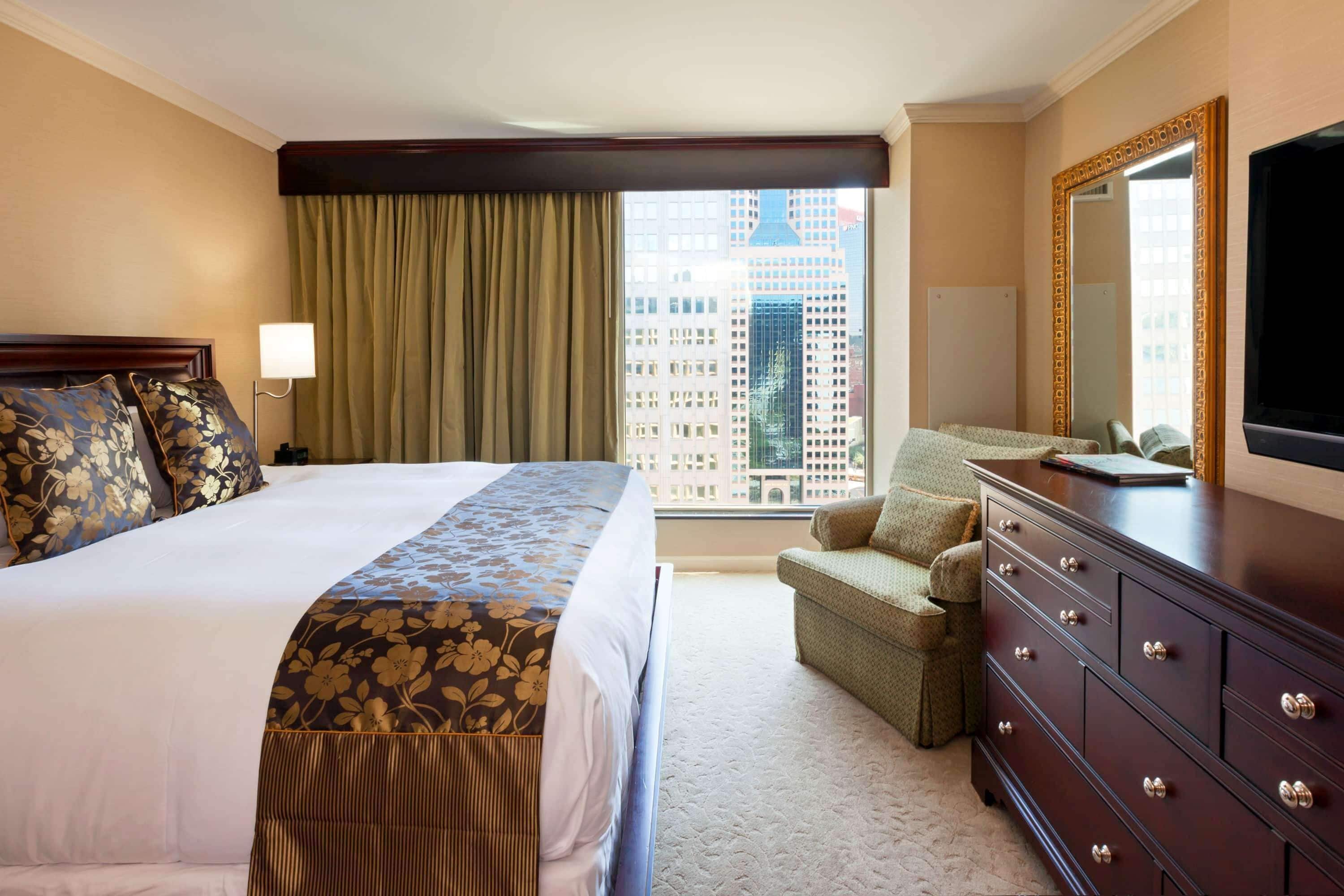 A hotel room with a large bed, a dresser, a chair, and a view of the cityscape through the window.