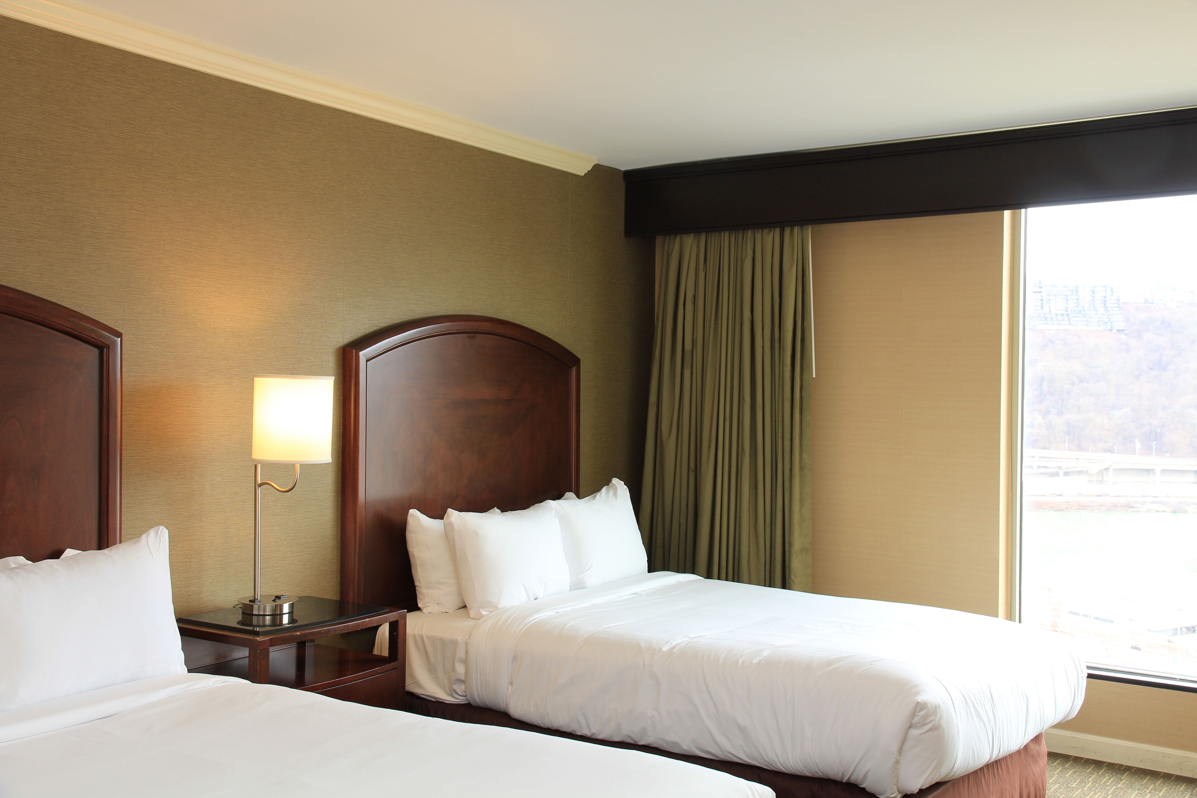 Een hotelkamer met twee bedden, een nachtkastje met lamp, een groot raam met gordijnen en uitzicht op een brug.
