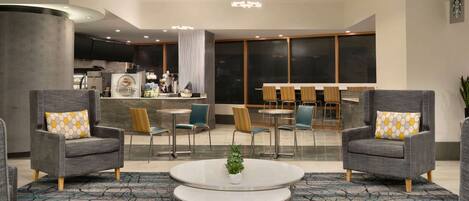 Un moderno vestíbulo de hotel con sillones grises, una cafetería y una zona de estar con mesas y sillas.