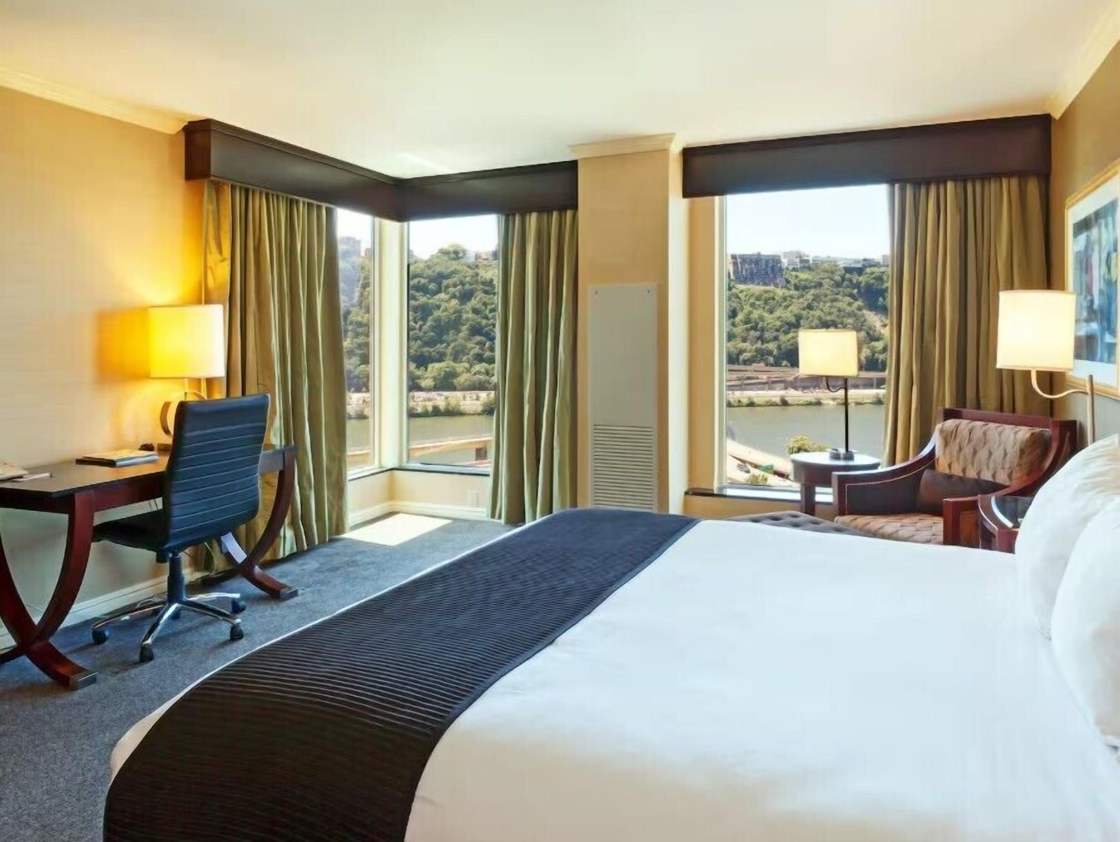 A hotel room with a large bed, a desk, a chair, a small table, and a view of a forested area through the windows.