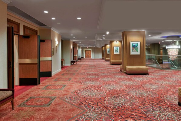 A hotel corridor with a red patterned carpet, wooden walls, and a staircase with glass railings.