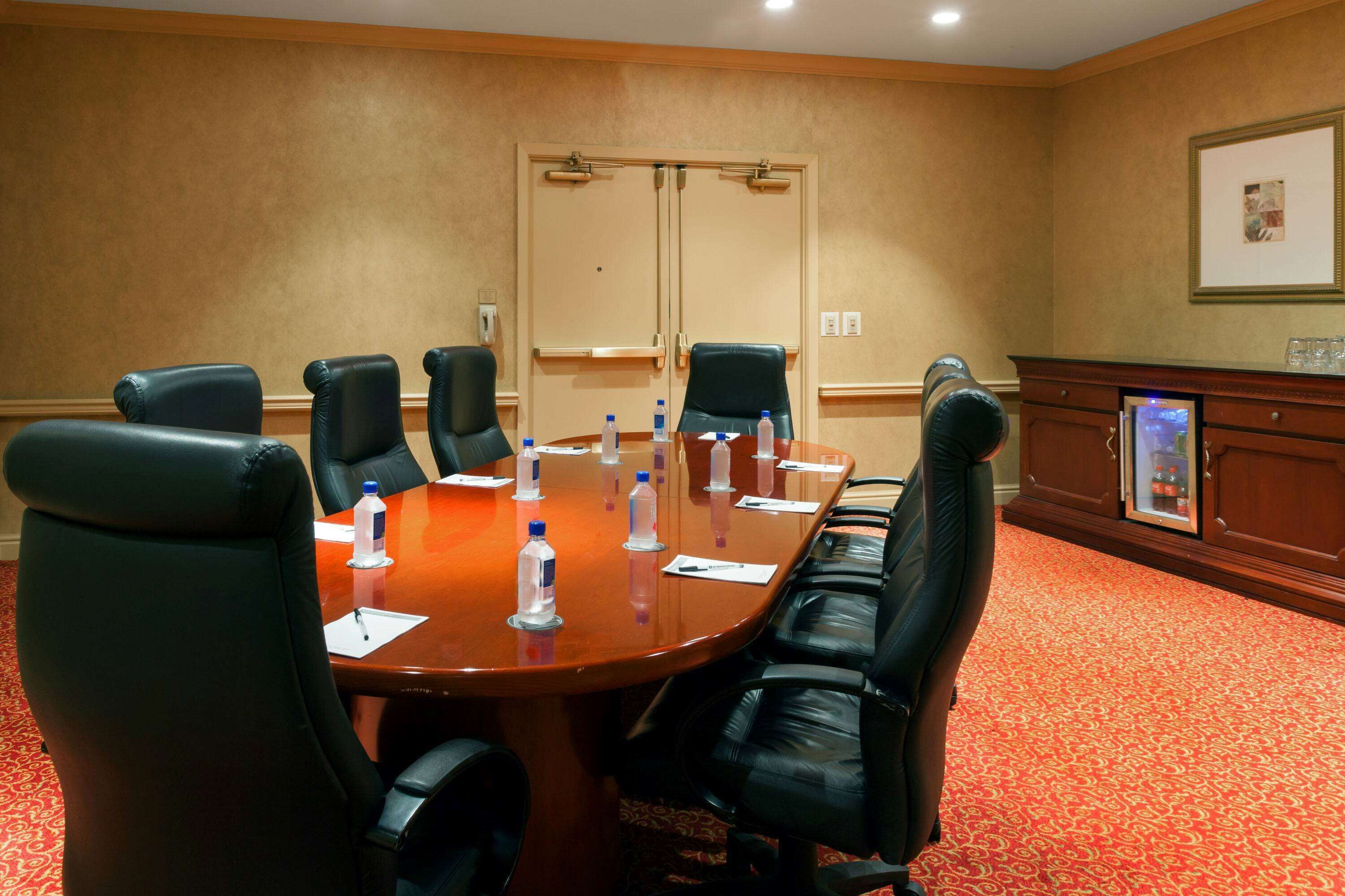A conference room with a wooden table, black leather chairs, water bottles, and a red carpeted floor.
