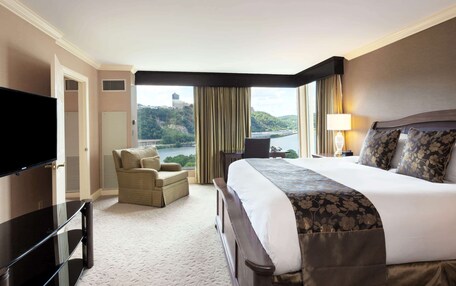 A hotel room with a large bed, a TV, a chair, a desk, and a view of a river and hills.