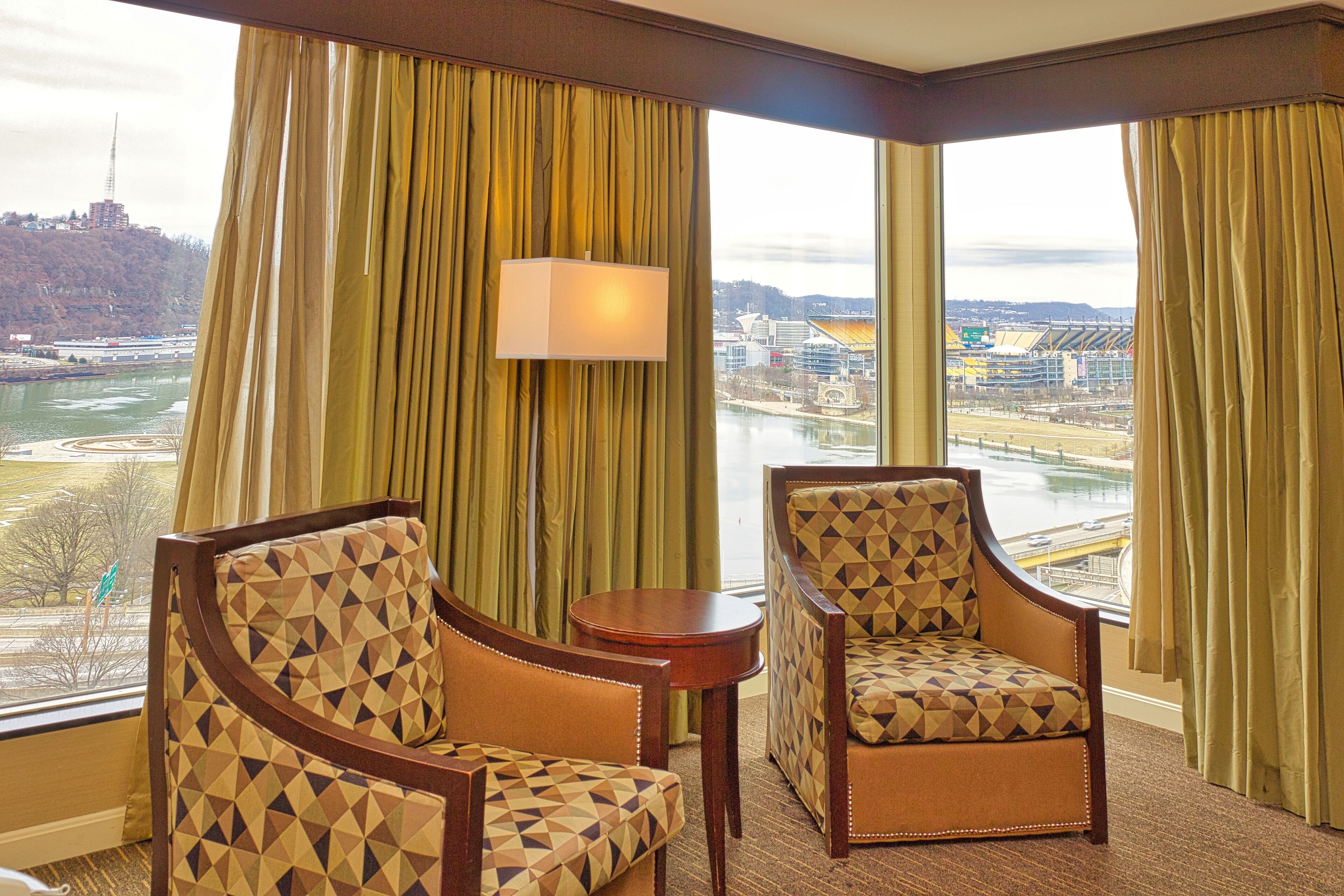 A hotel room with two armchairs, a small table, and a view of a river and cityscape through large windows.