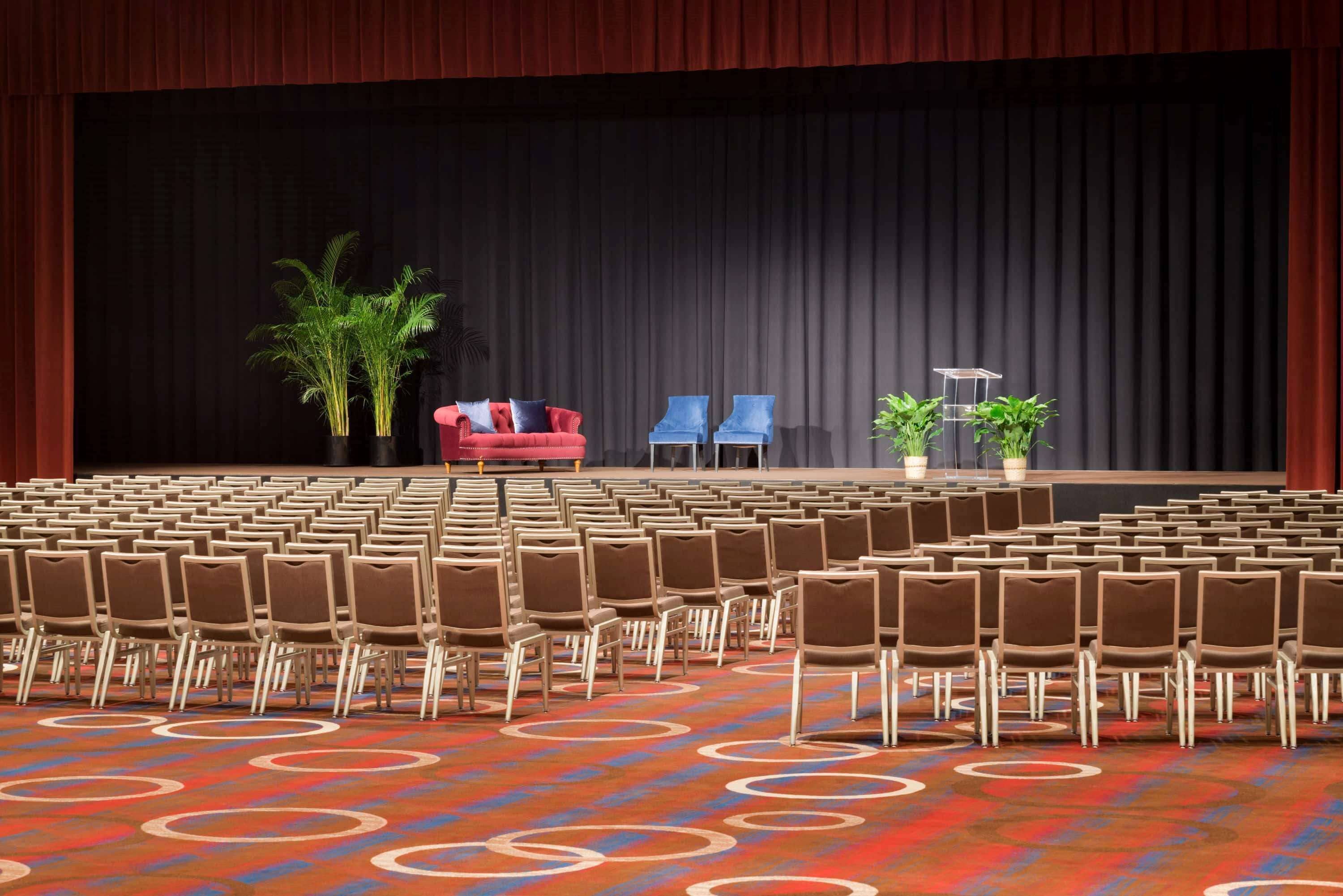Une salle de conférence avec des rangées de chaises, une scène dotée d’un fond noir, un canapé rouge et deux plantes en pot.