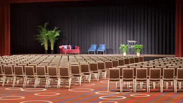 Une salle de conférence avec des rangées de chaises, une scène dotée d’un fond noir, un canapé rouge et deux plantes en pot.