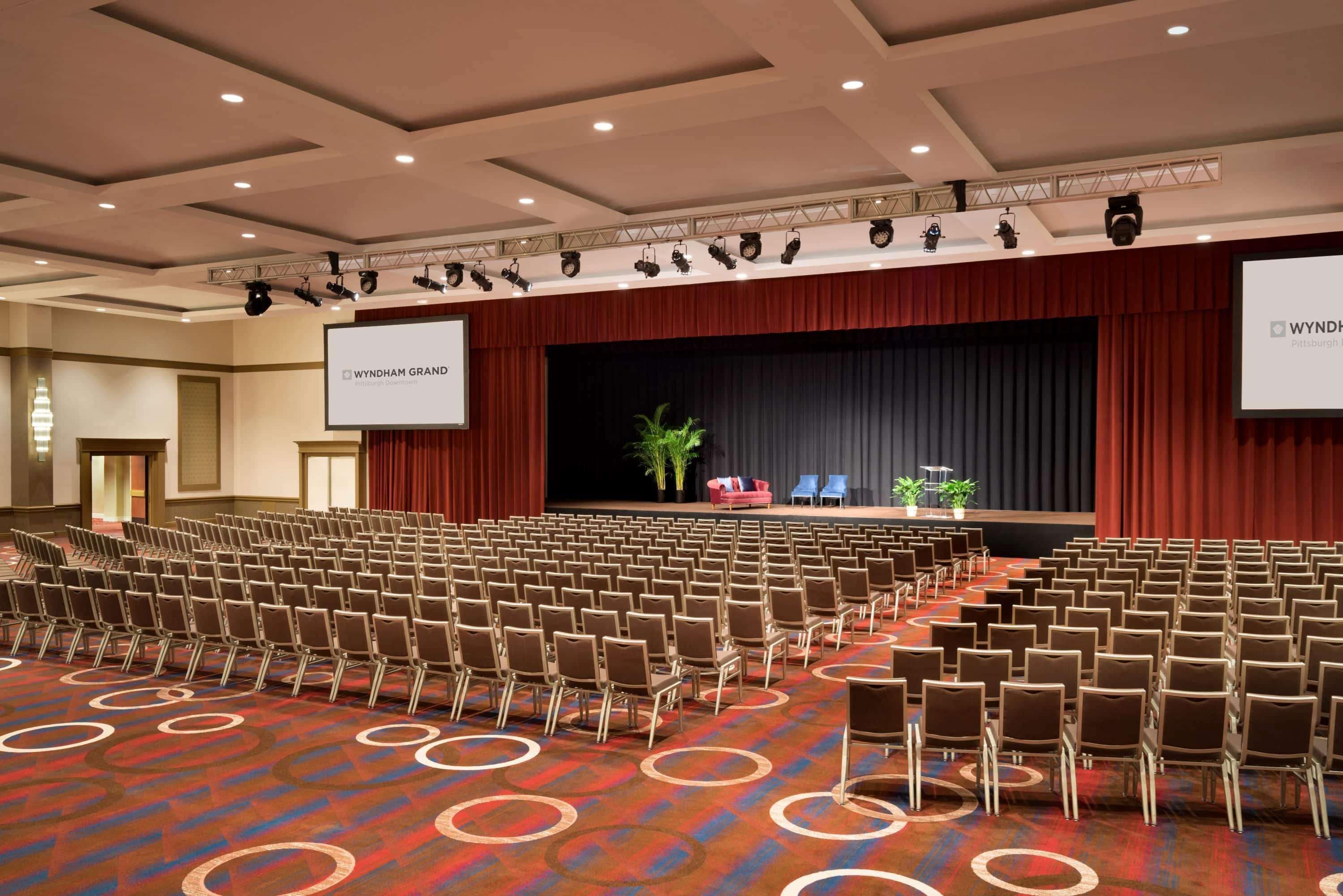 Une salle de conférence avec des rangées de chaises, une scène dotée d’un écran et un tapis orné de motifs circulaires.