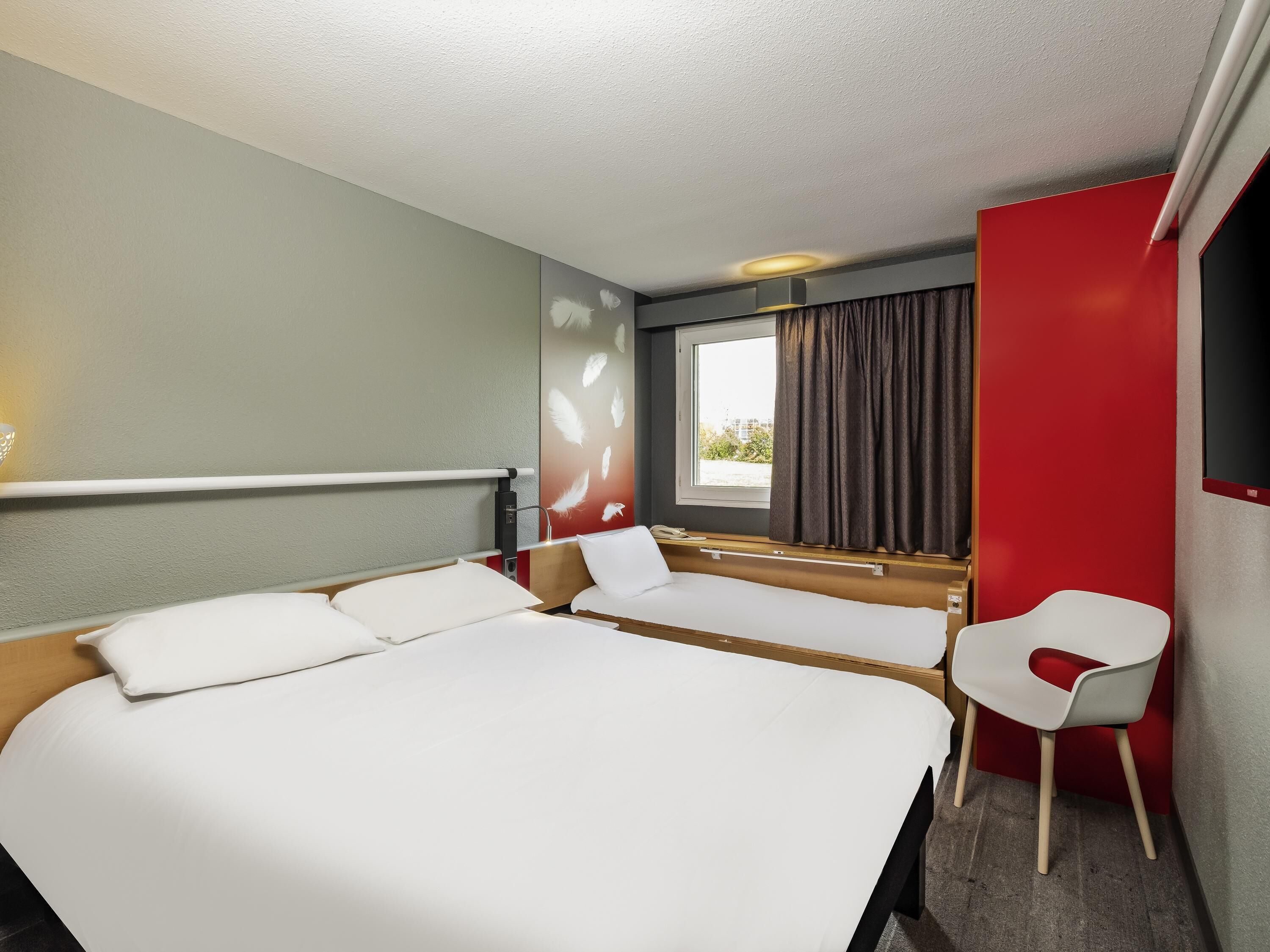Ein Hotelzimmer mit einem großen Bett, einem Nachttisch, einem Sessel, einem Fenster mit Vorhängen und einem an der Wand befestigten Fernseher.