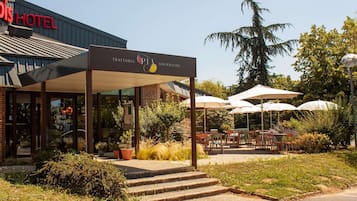 L’extérieur d’un hôtel, avec un espace extérieur couvert aménagé en coin salon, des marches menant à l’entrée et une enseigne indiquant un restaurant appelé « Trattoria PIA Gourmet ».