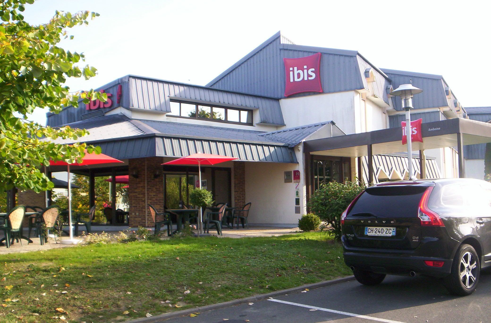 A modern building with a prominent 'ibis' sign, outdoor seating area, and a parked car.