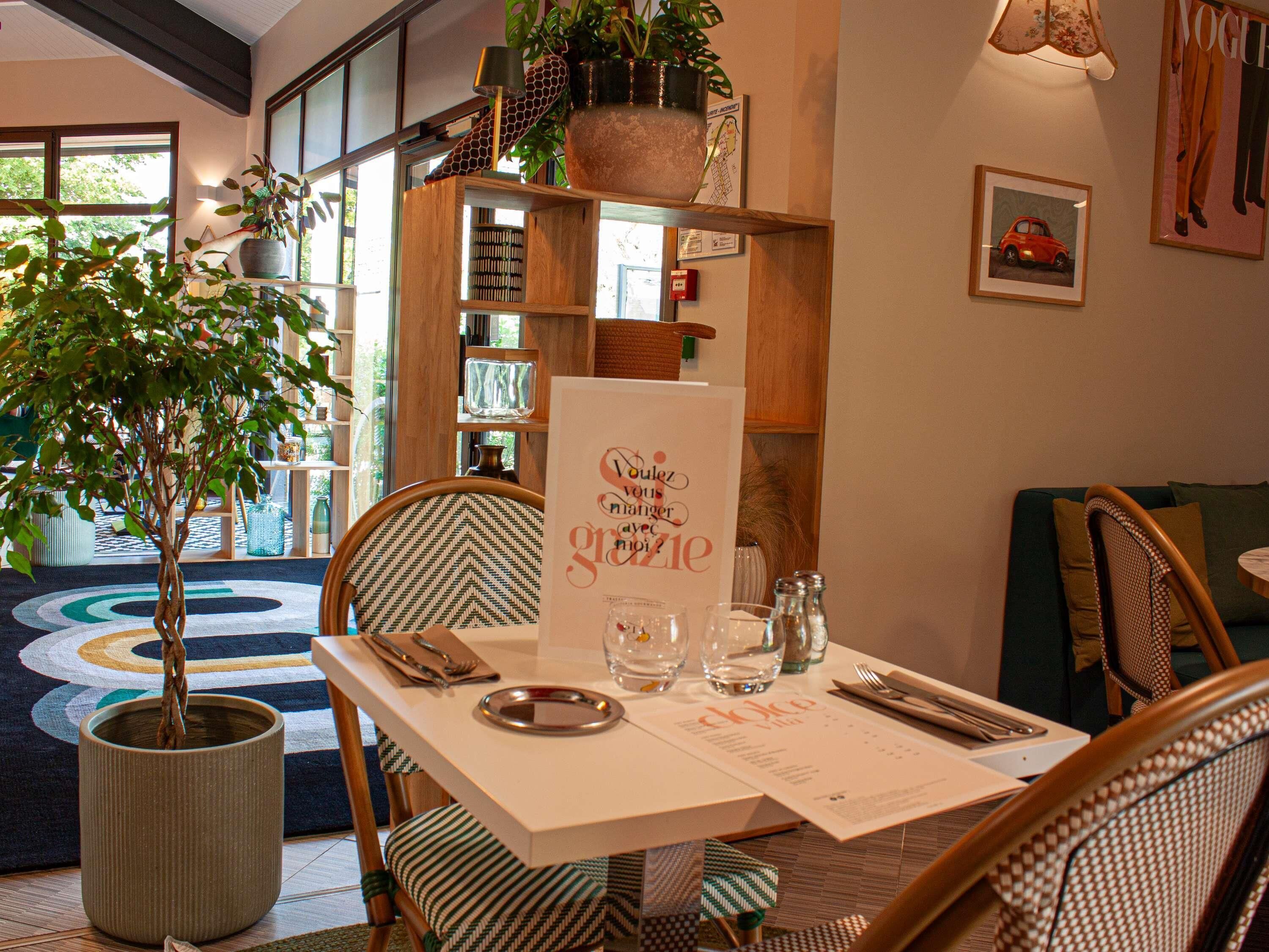 A dining area with a table set for two, surrounded by wicker chairs, a potted plant, and a bookshelf with various items.