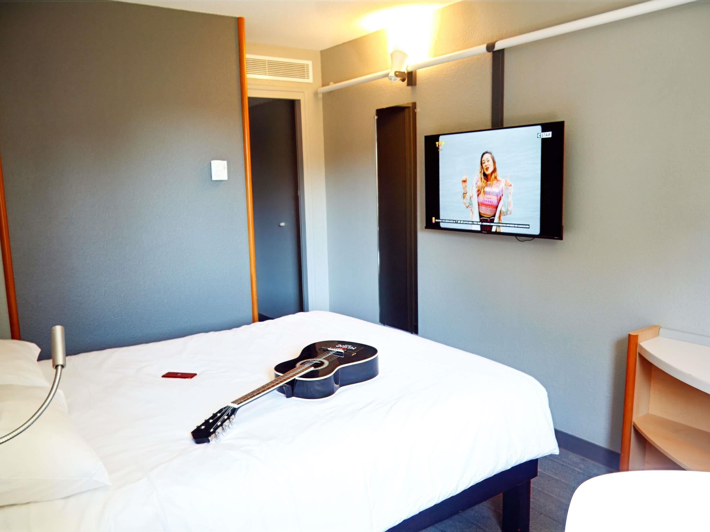 A hotel room with a bed, a guitar, a TV, and a bedside table.
