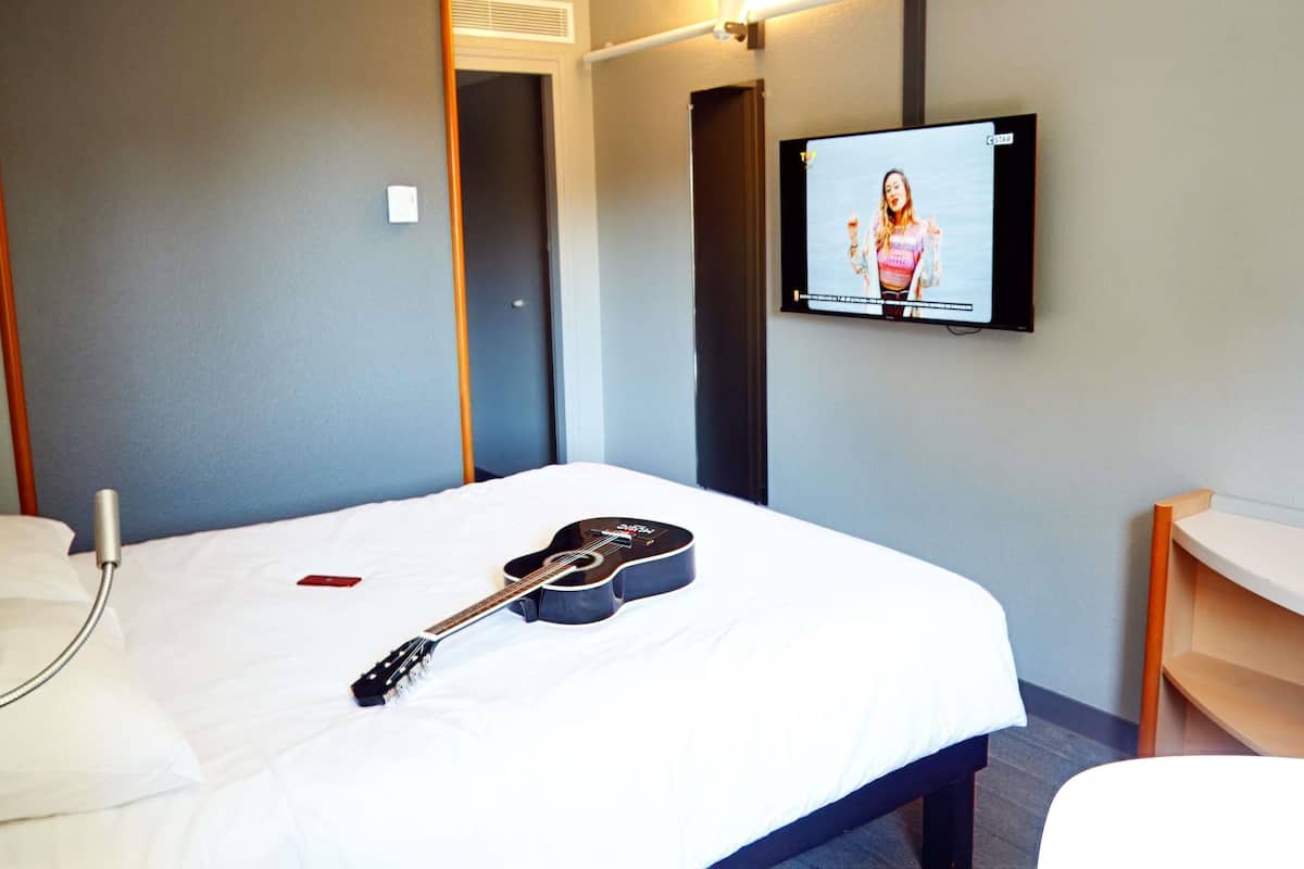 A hotel room with a bed, a guitar, a TV, and a bedside table.