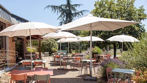 An outdoor patio area with tables and chairs under large umbrellas, surrounded by gravel and greenery. - Ibis Amboise (Amboise)