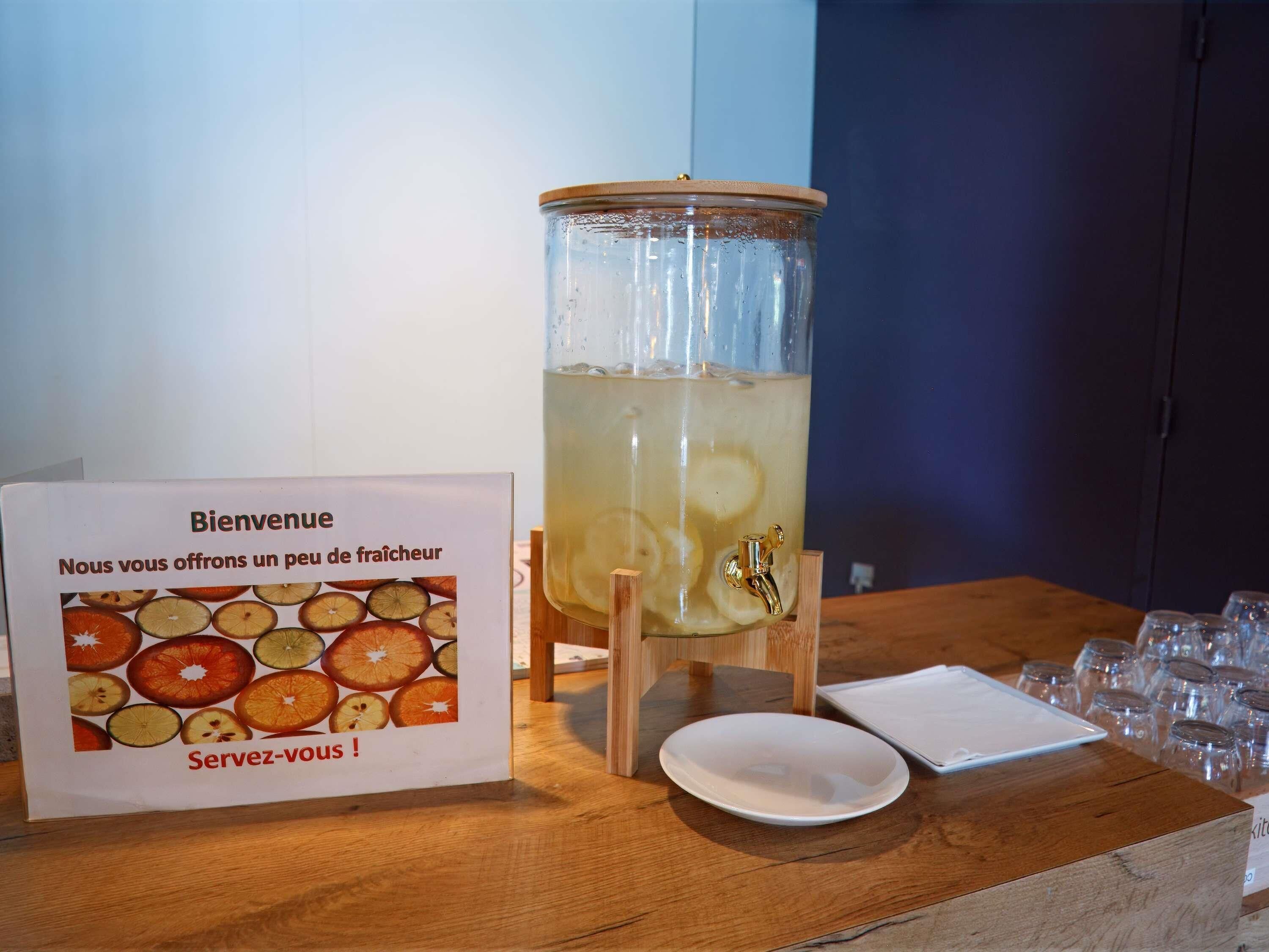 A welcome sign with fruit illustrations, a large beverage dispenser, and glasses on a wooden table.