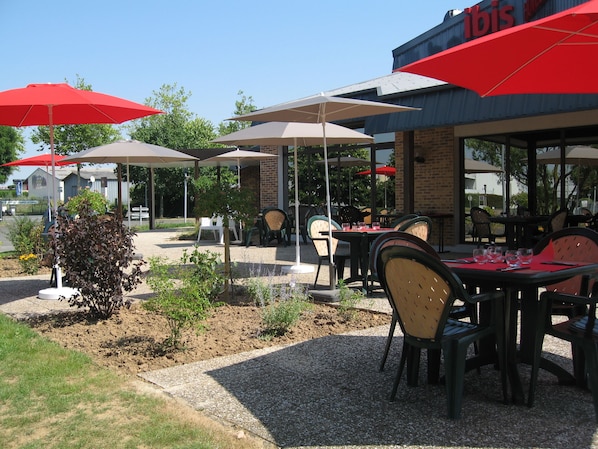 An outdoor seating area with tables and chairs under umbrellas, a brick building in the background, and a gravel path. - Ibis Amboise (Amboise)