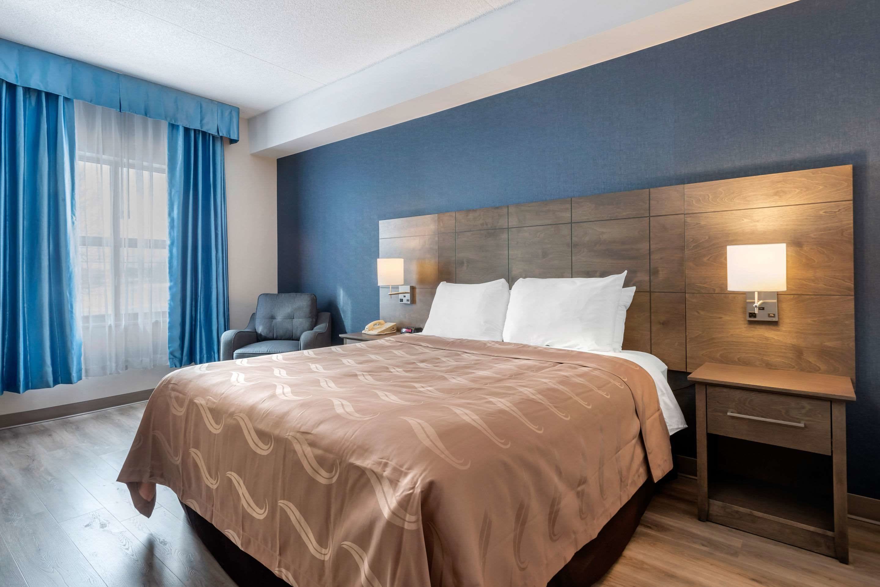 A hotel room with a large bed, two bedside lamps, a chair, a nightstand, and a window with blue curtains.