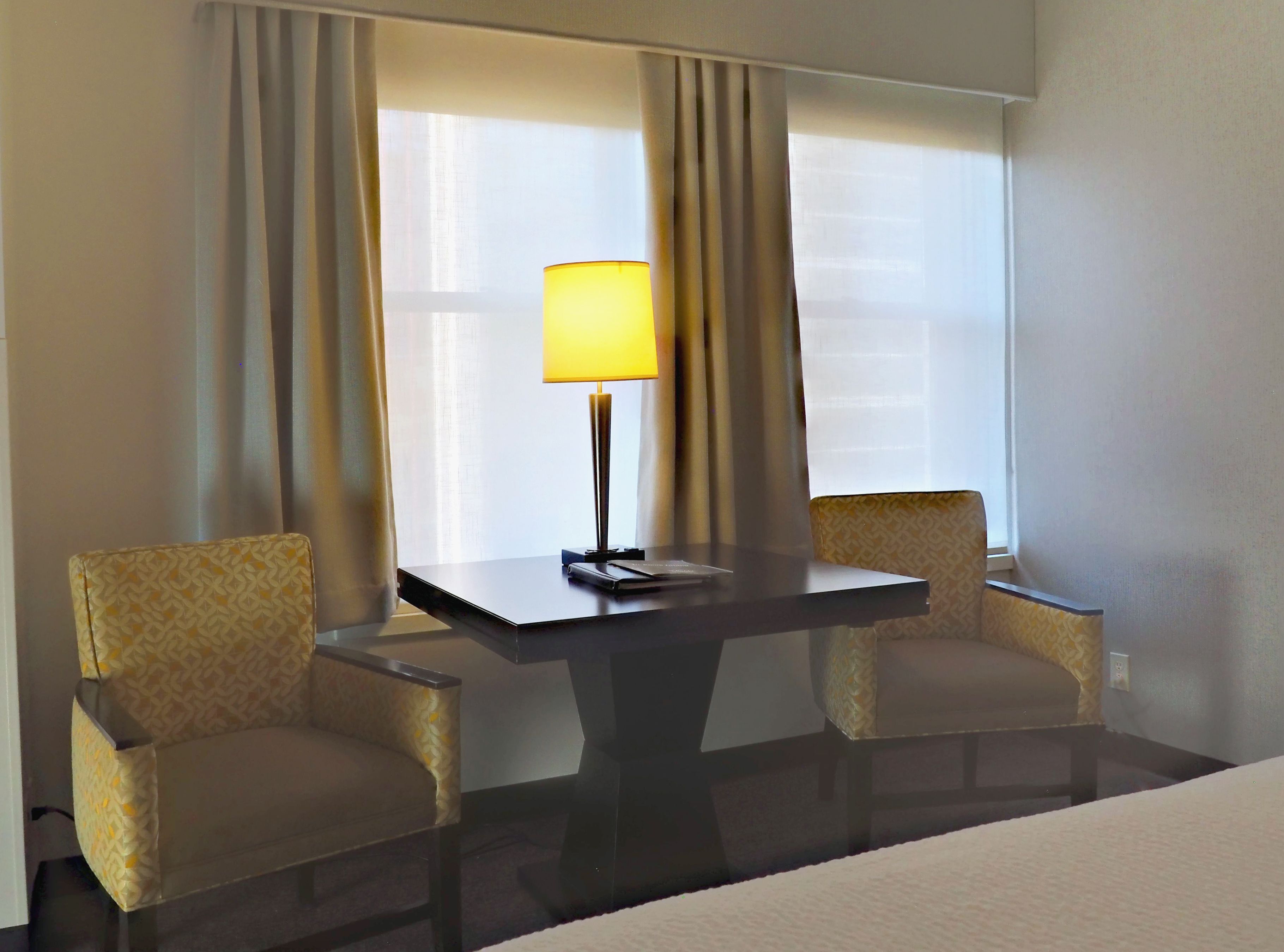 A hotel room with a bed, a table with a lamp, and two patterned armchairs.