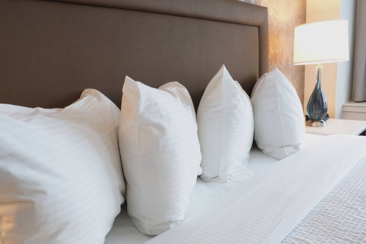 A neatly made bed with white linens and pillows, a brown headboard, and a bedside lamp.