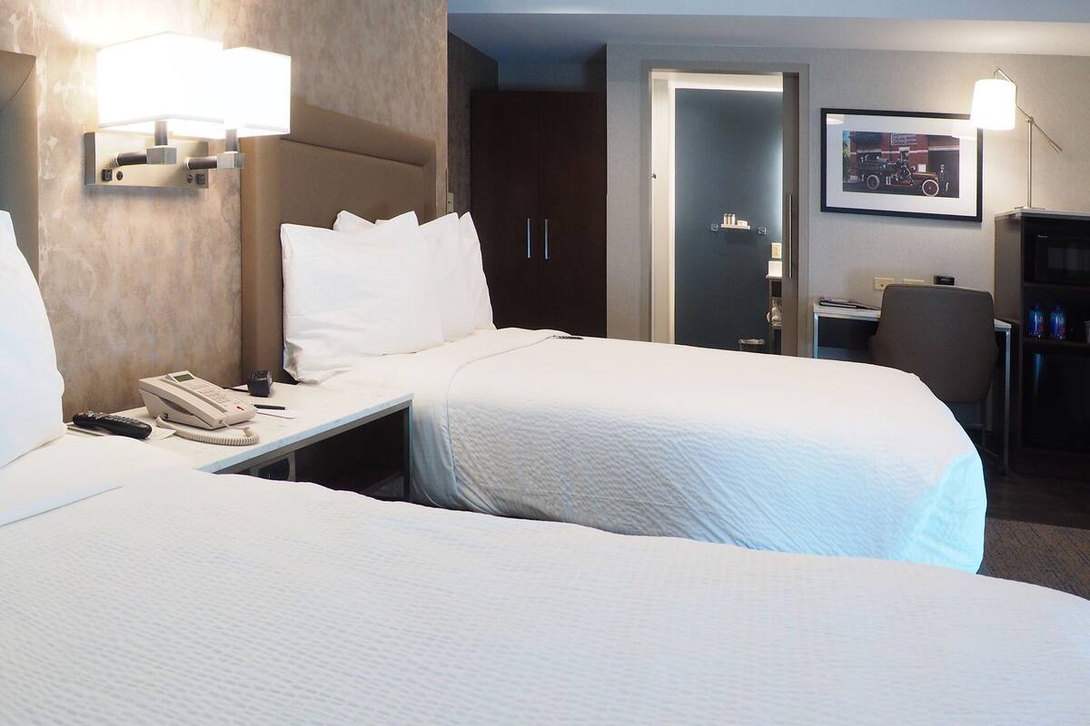 A hotel room with two beds, a bedside table with a phone, a lamp, and a framed picture on the wall.