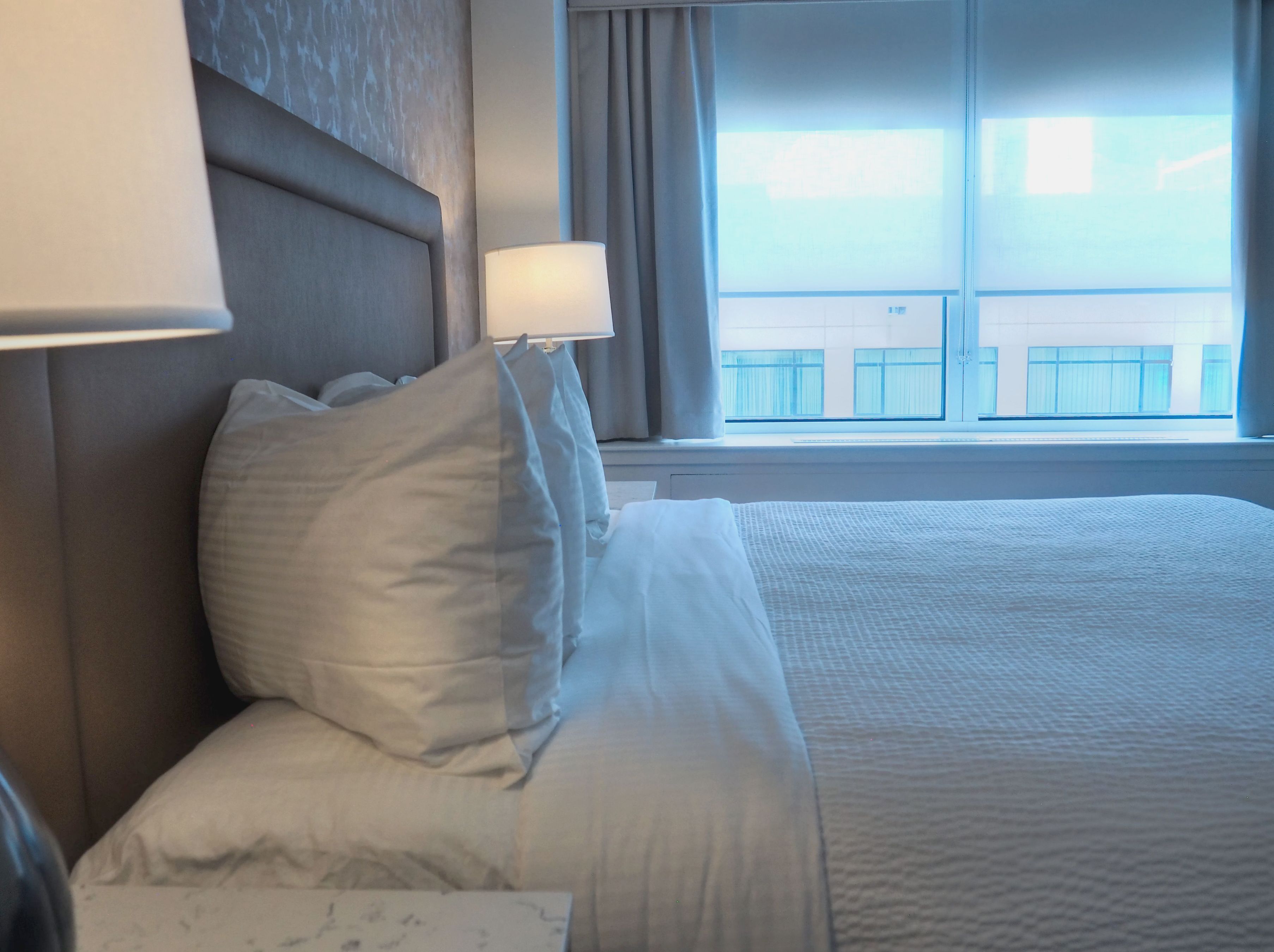 A hotel room with a bed, a nightstand, a lamp, and a window with a view of buildings.